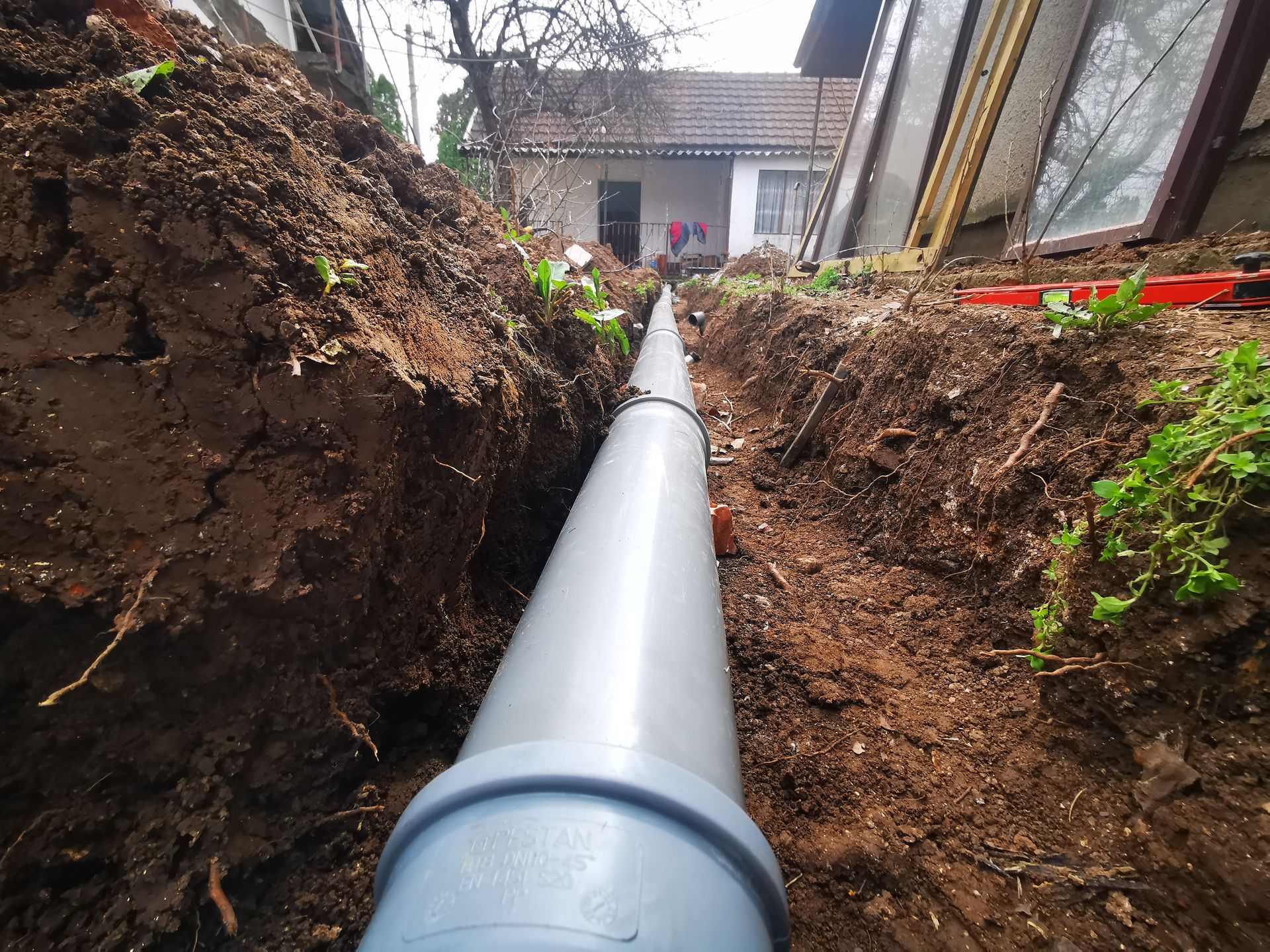 Large gray pipe laid in a narrow trench between dirt banks near a house.