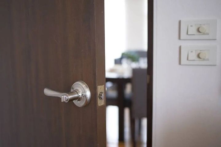 Porta di legno parzialmente aperta con maniglia argentata, che si apre su una luminosa stanza interna con interruttori della luce a parete.