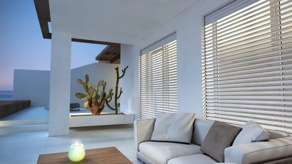 Patio moderno con divano bianco, tavolo in legno, tende e cactus in vaso al tramonto.