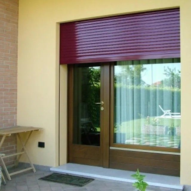 Patio con porte a vetri marroni, tenda a rullo viola e tavolino pieghevole accanto a un prato.