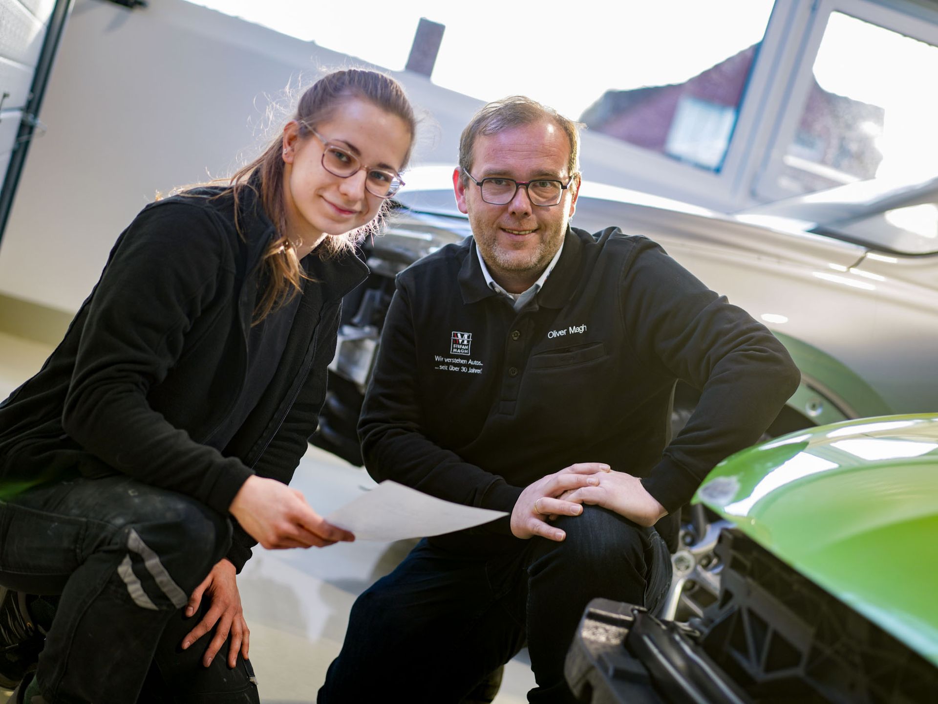 Geschäftsführer Oliver Magh und Mitarbeiterin Lenina sitzen neben einem grünen Auto.