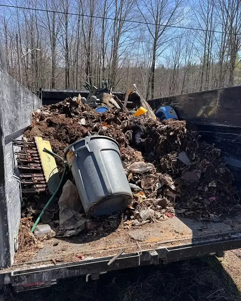 Dumpster filled with yard debris and trash, including a tipped gray bin in an outdoor wooded area.