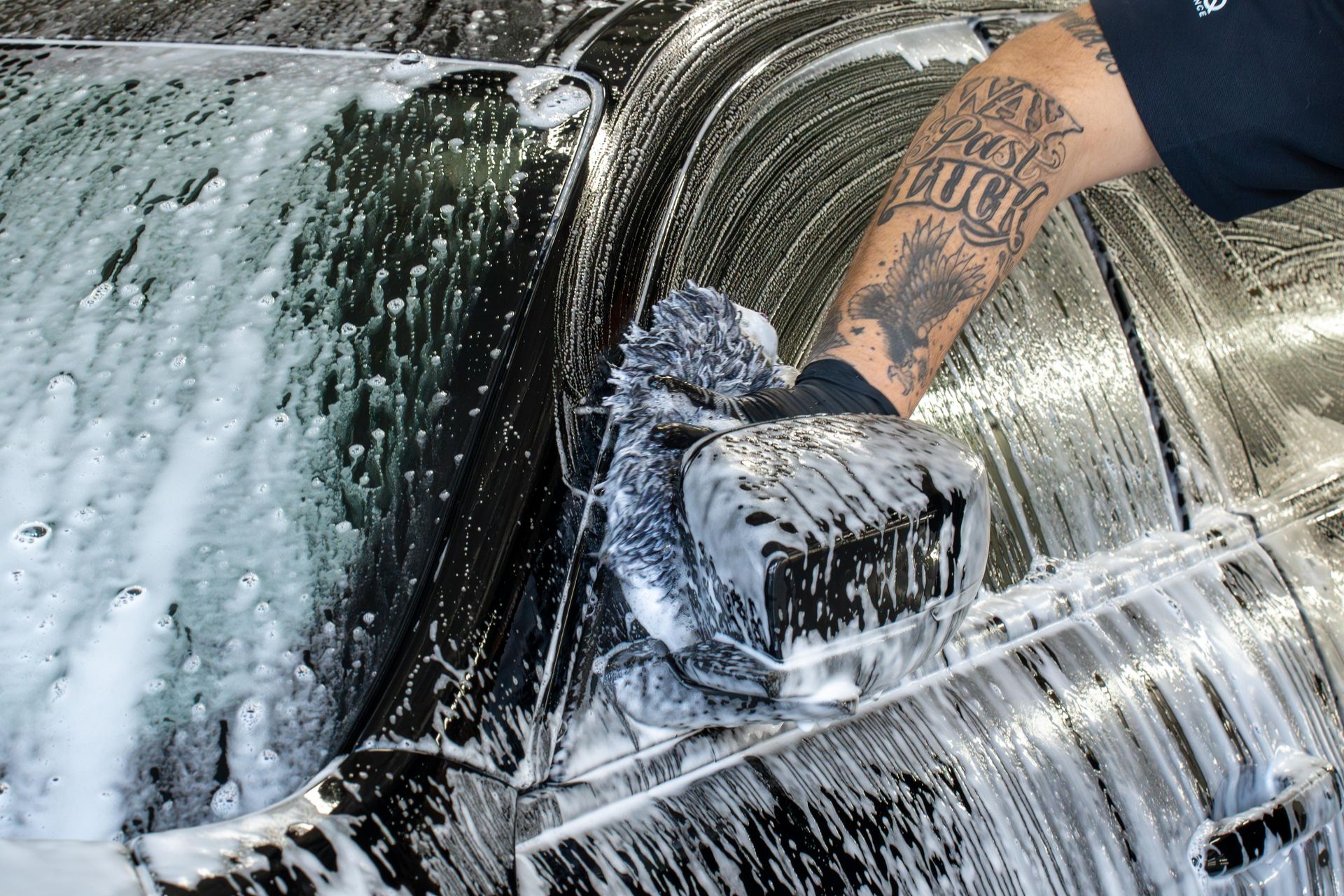 Person washing a car with a soapy mitt; arm with tattoo visible.