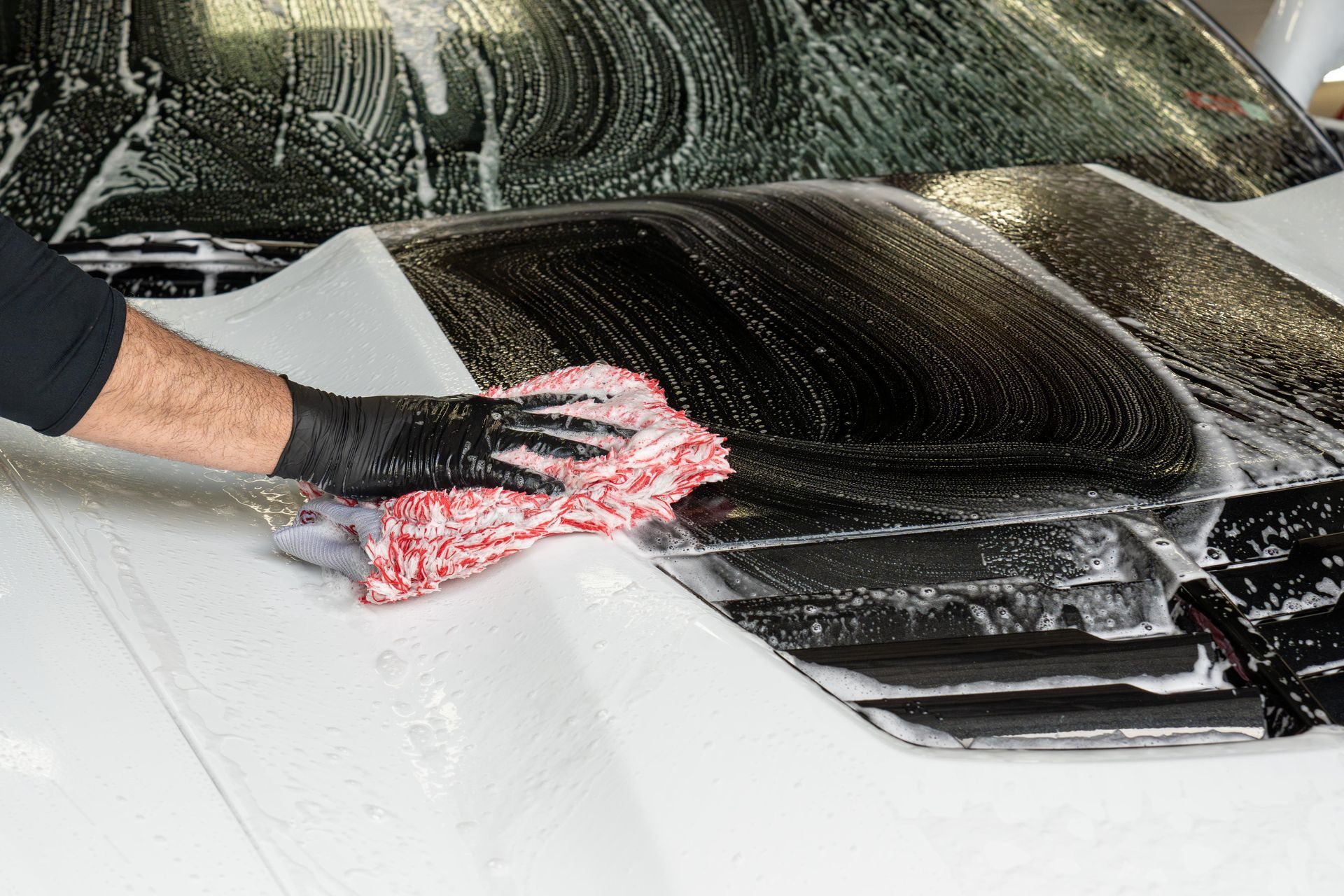 Person washing a white car with a soapy cloth, wearing black gloves.