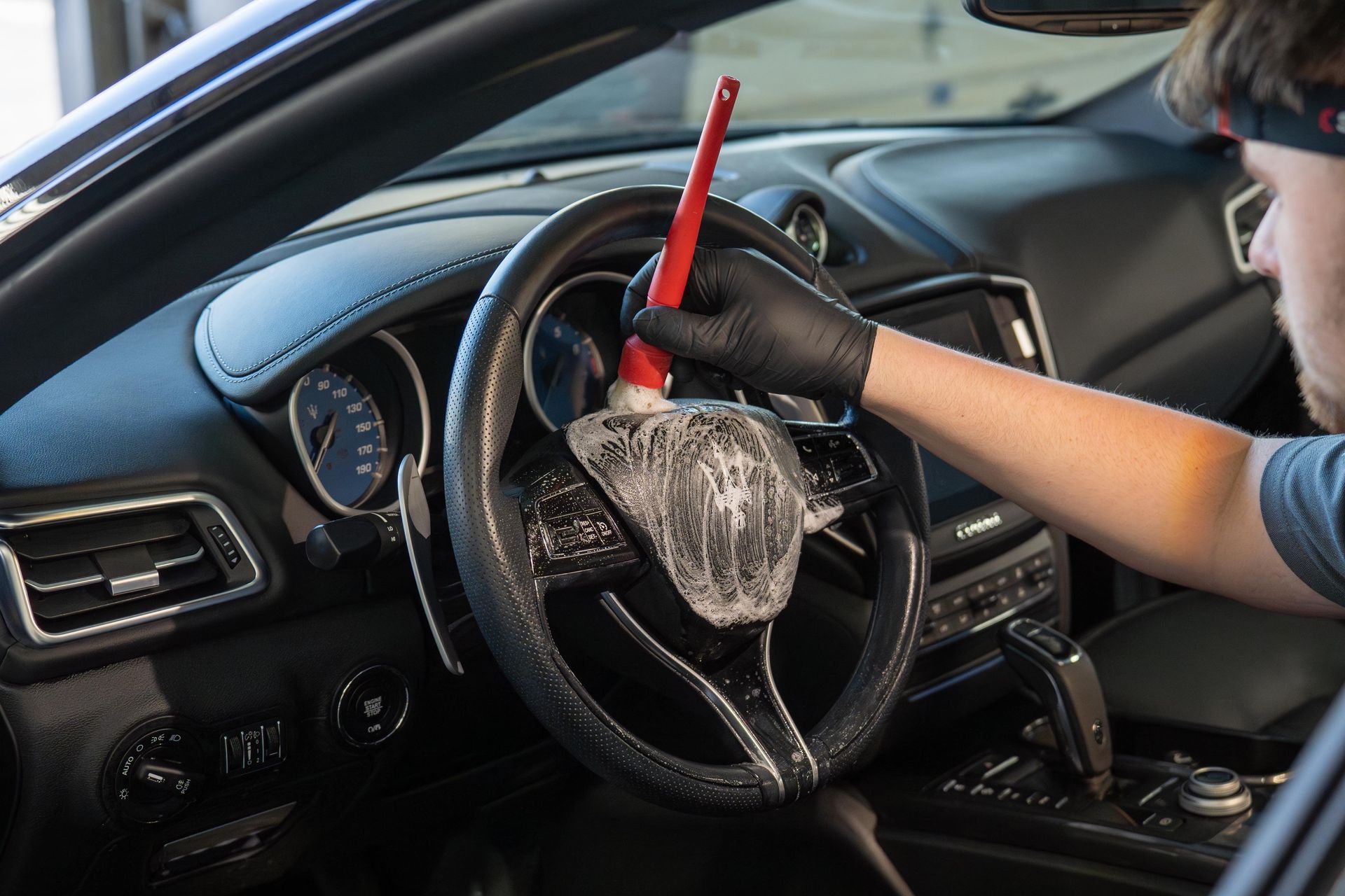 Person wearing black gloves cleaning a car's steering wheel with a brush and soapy solution.