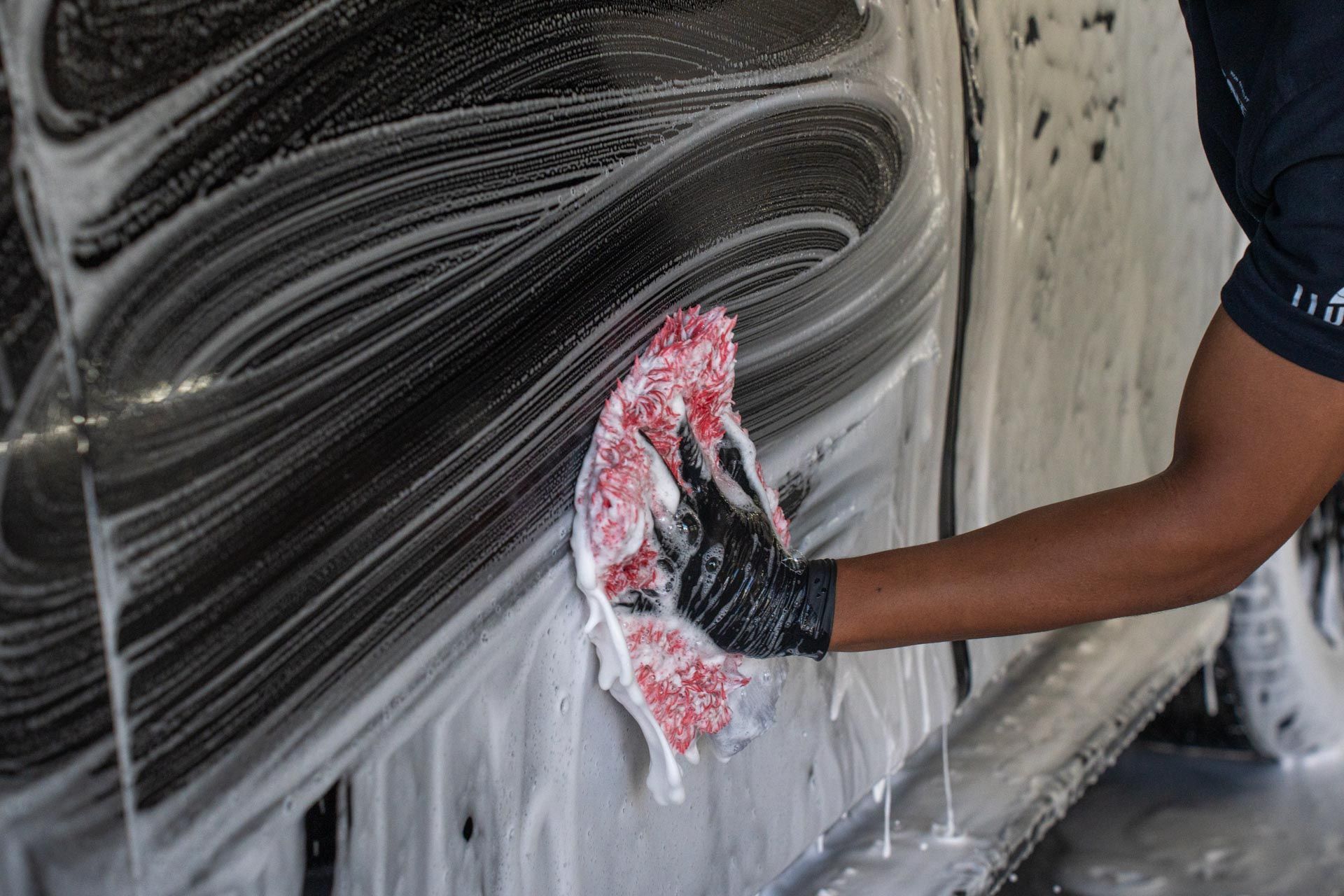 Person washing a black vehicle with soapy water, using a red and white wash mitt.