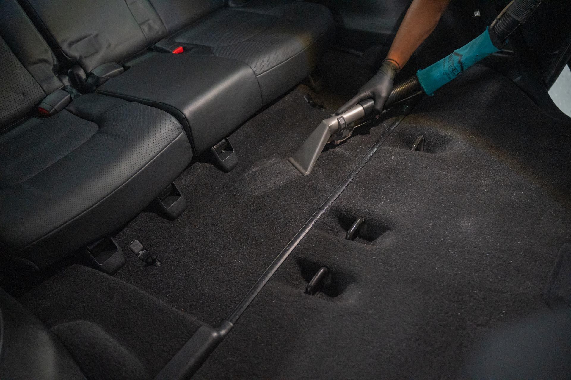 Person vacuuming a car's interior carpet, black seats visible. They are wearing gloves and using a blue vacuum.