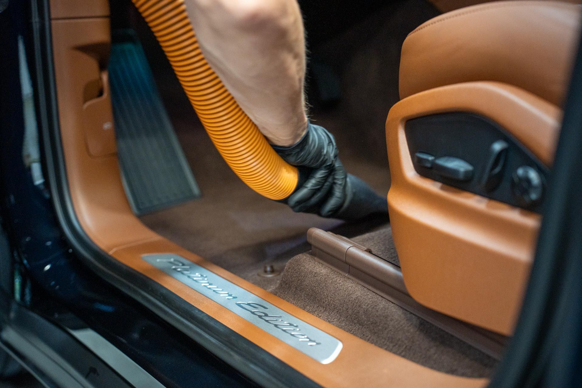 Person vacuuming the interior of a car, focusing on the carpet and door sill with an orange hose.