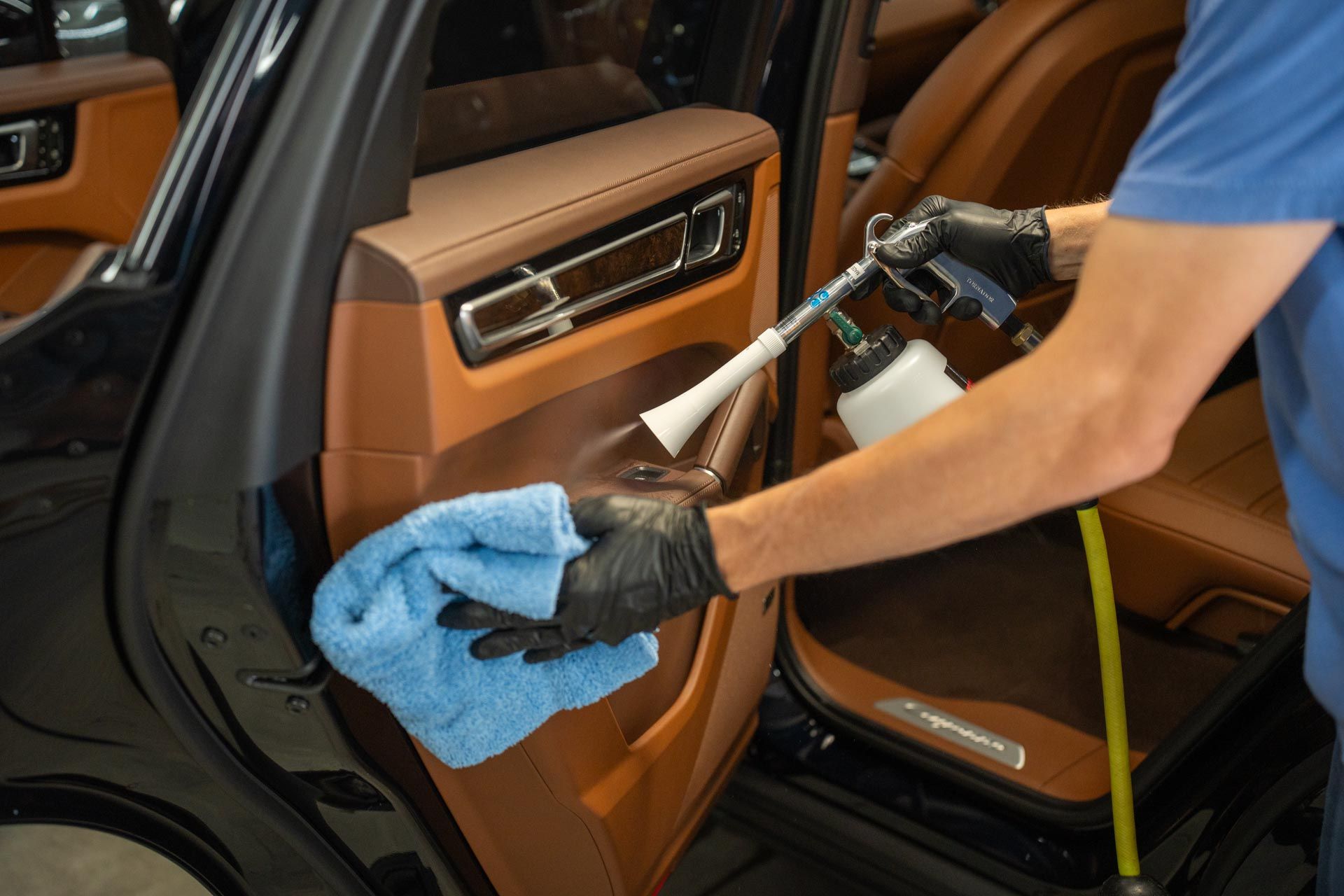 Person cleaning a car's interior door panel with a spray gun and microfiber cloth; tan leather and black car.