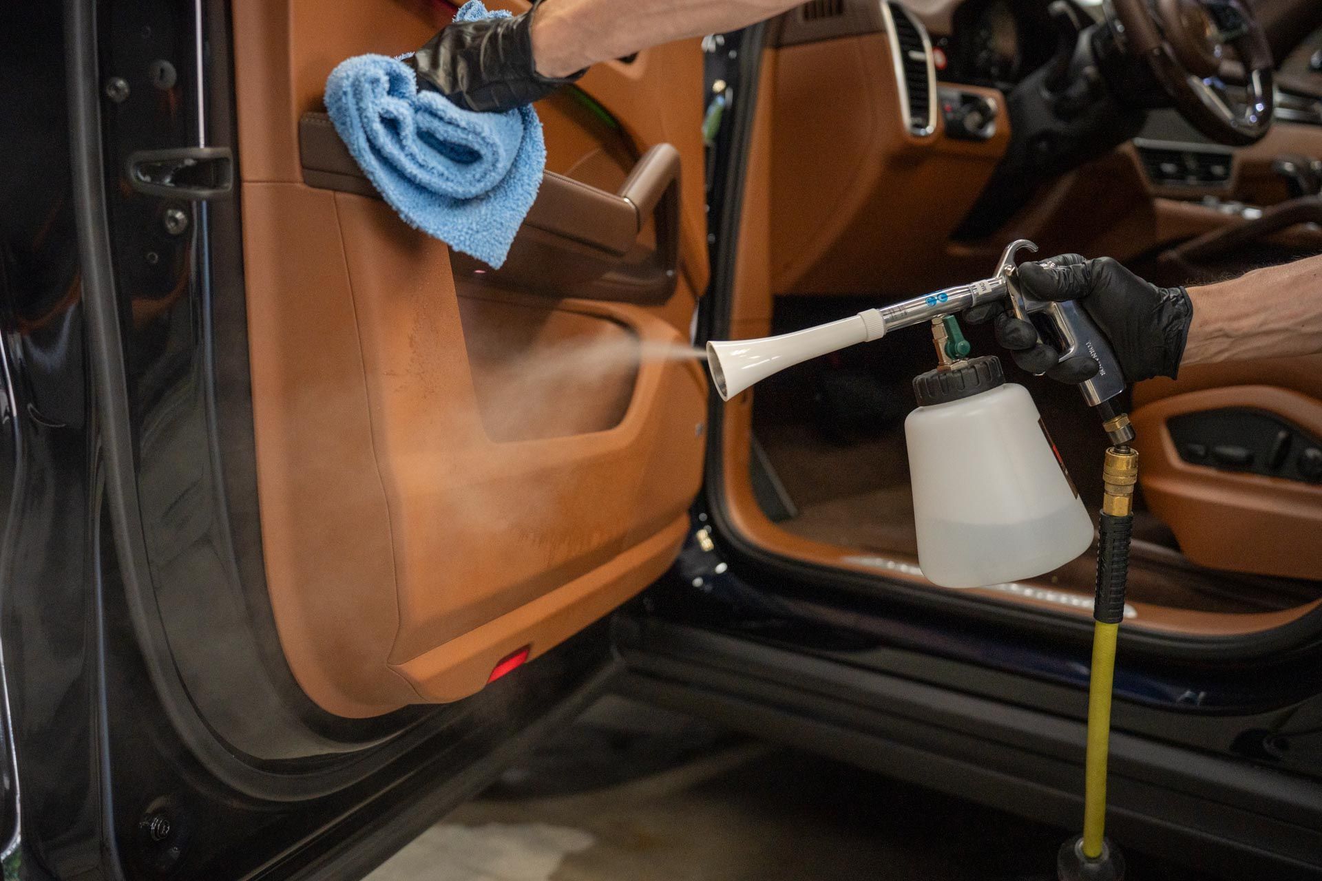 Person cleaning car door panel with a spray tool and cloth. Brown leather interior.