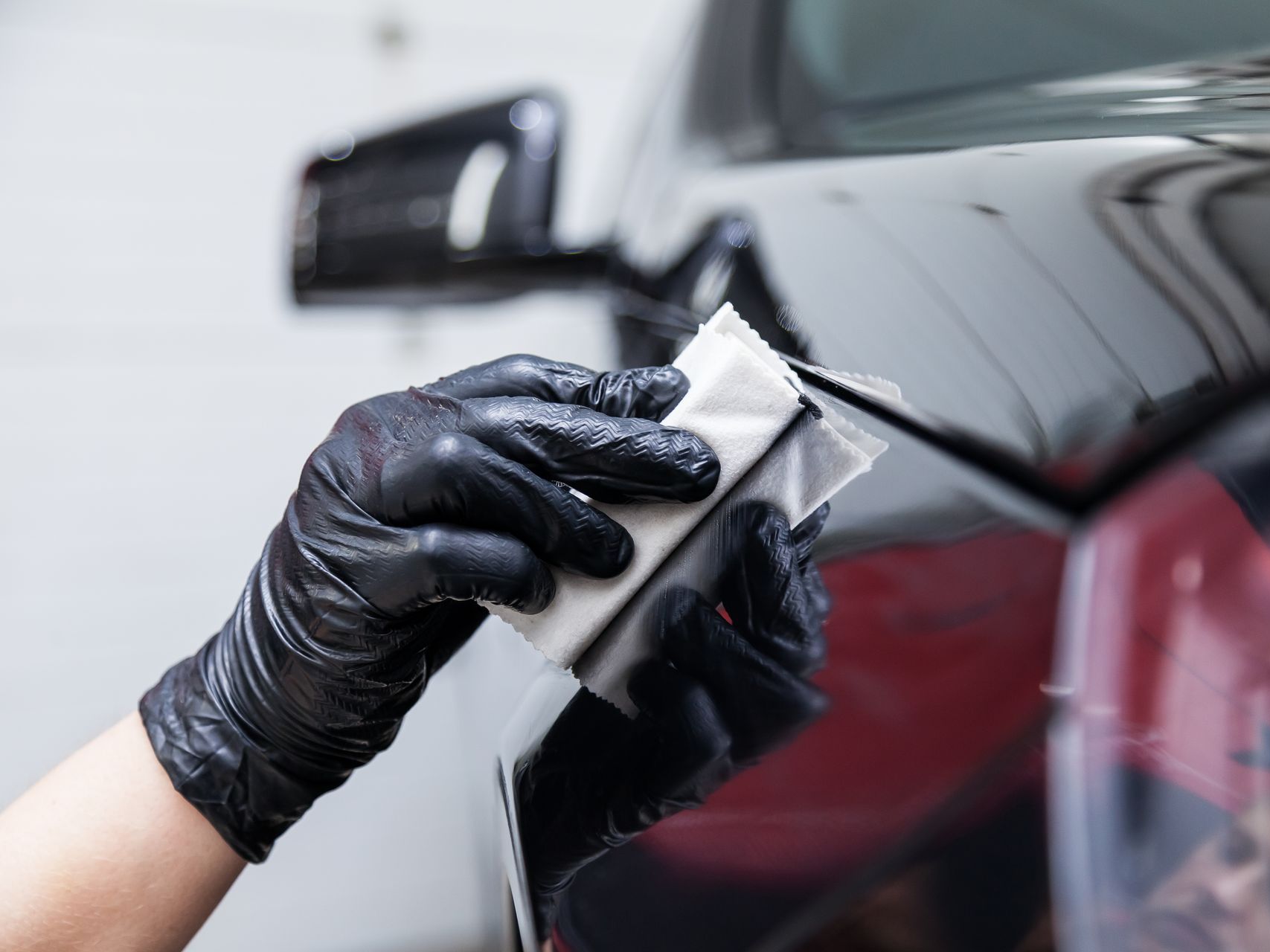 Gloved hands applying detailing product to a black car's body, near a side mirror and taillight.