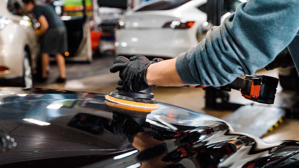 Person in black gloves polishing a car's black hood with a rotary tool in a car workshop.