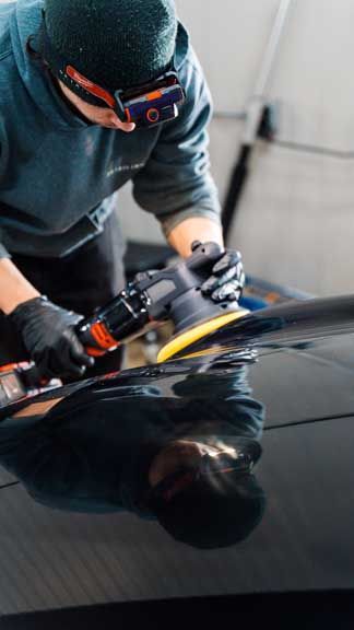 Person polishing a black car, wearing goggles and gloves, in a garage.