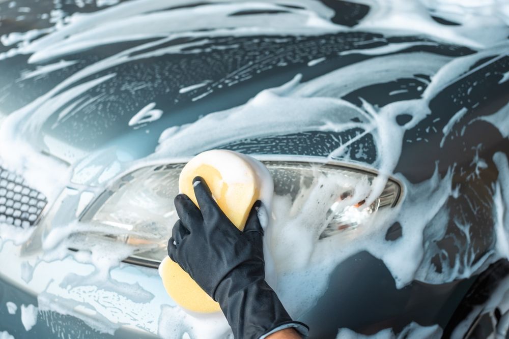Person wearing black glove washing car headlight with a sponge covered in soap suds.