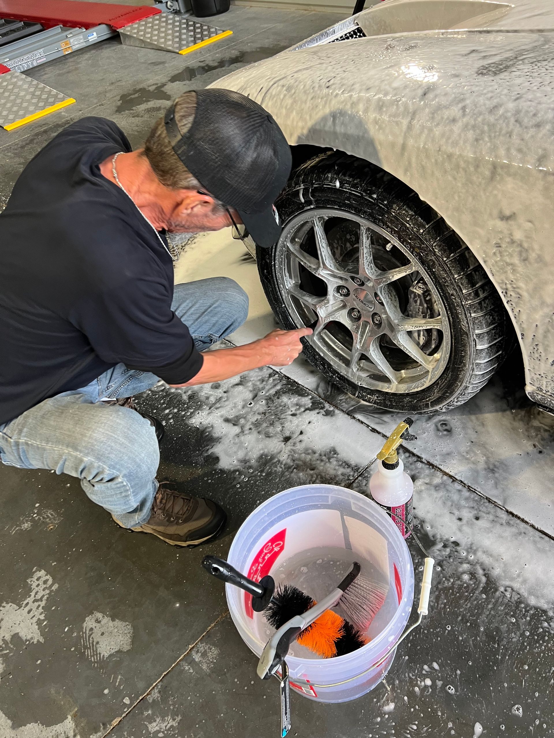 Man washes a car wheel with soapy water; bucket with tools sits nearby.