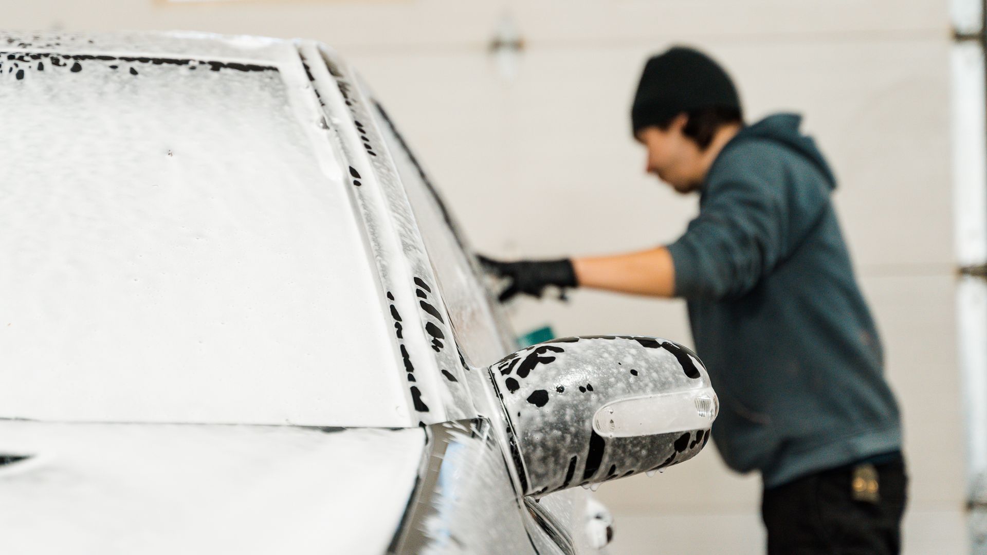 Person washing a black car covered in white soap suds in a garage.