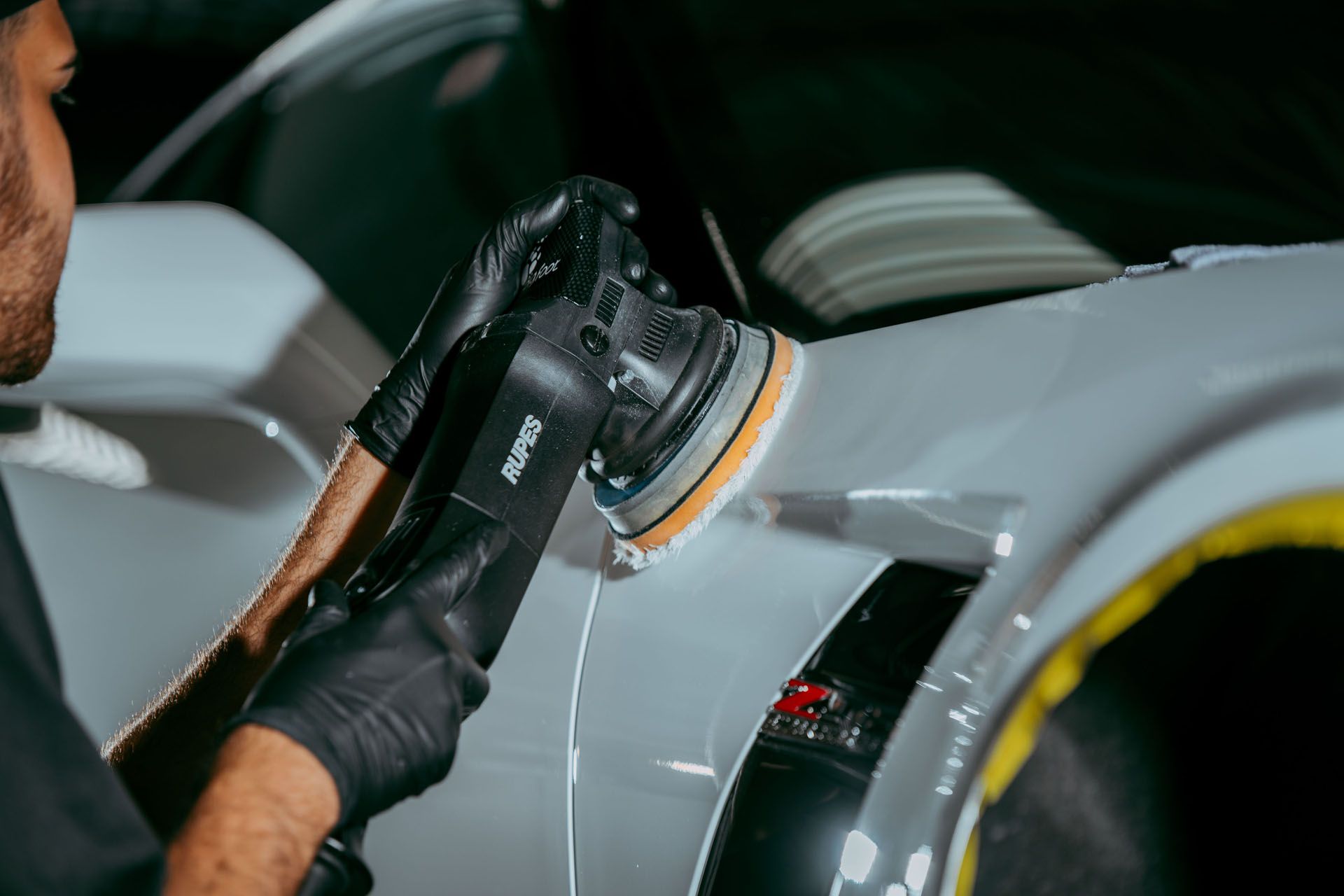 Person polishing a white car with an electric polisher in a garage.