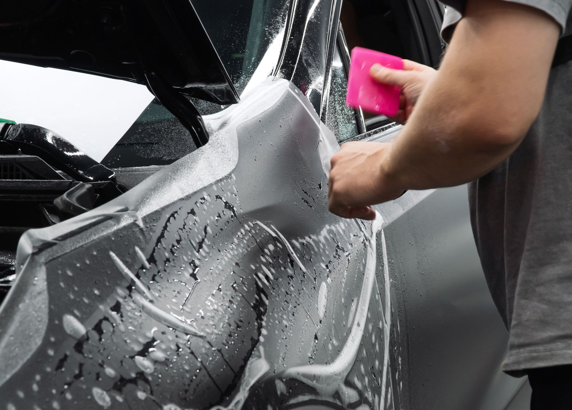 Gloved hand applying a coating to a shiny black car. The setting is outdoors with sunlight.