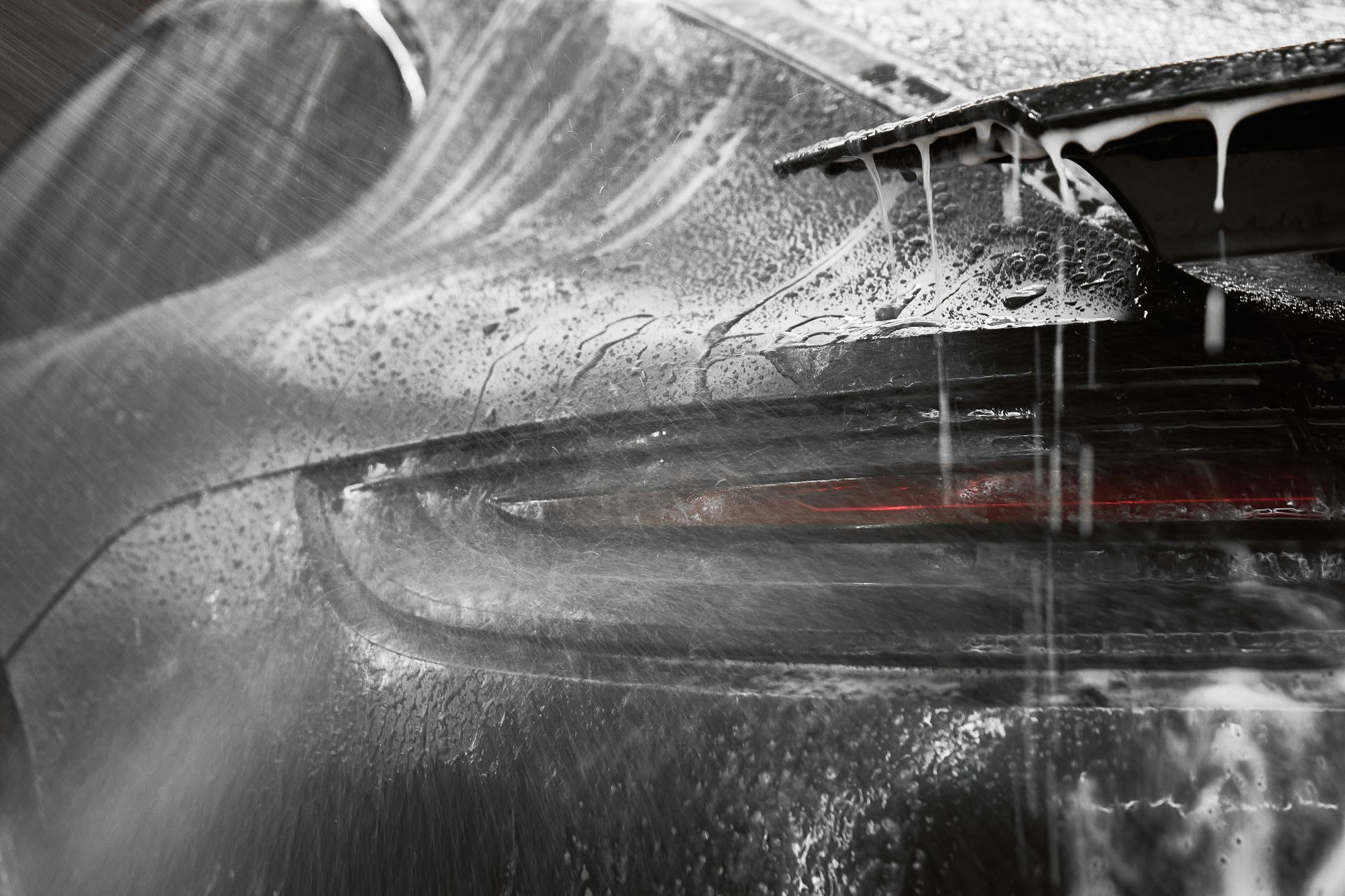 Car's rear end covered in soap suds during a wash; taillight visible, water dripping down.