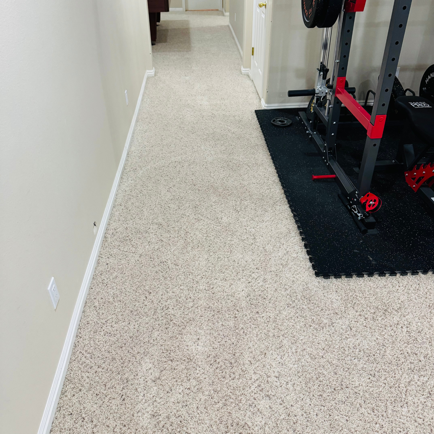 A hallway with light-colored speckled carpet, showing a doorway at the end and a home gym rack on a black rubber mat.