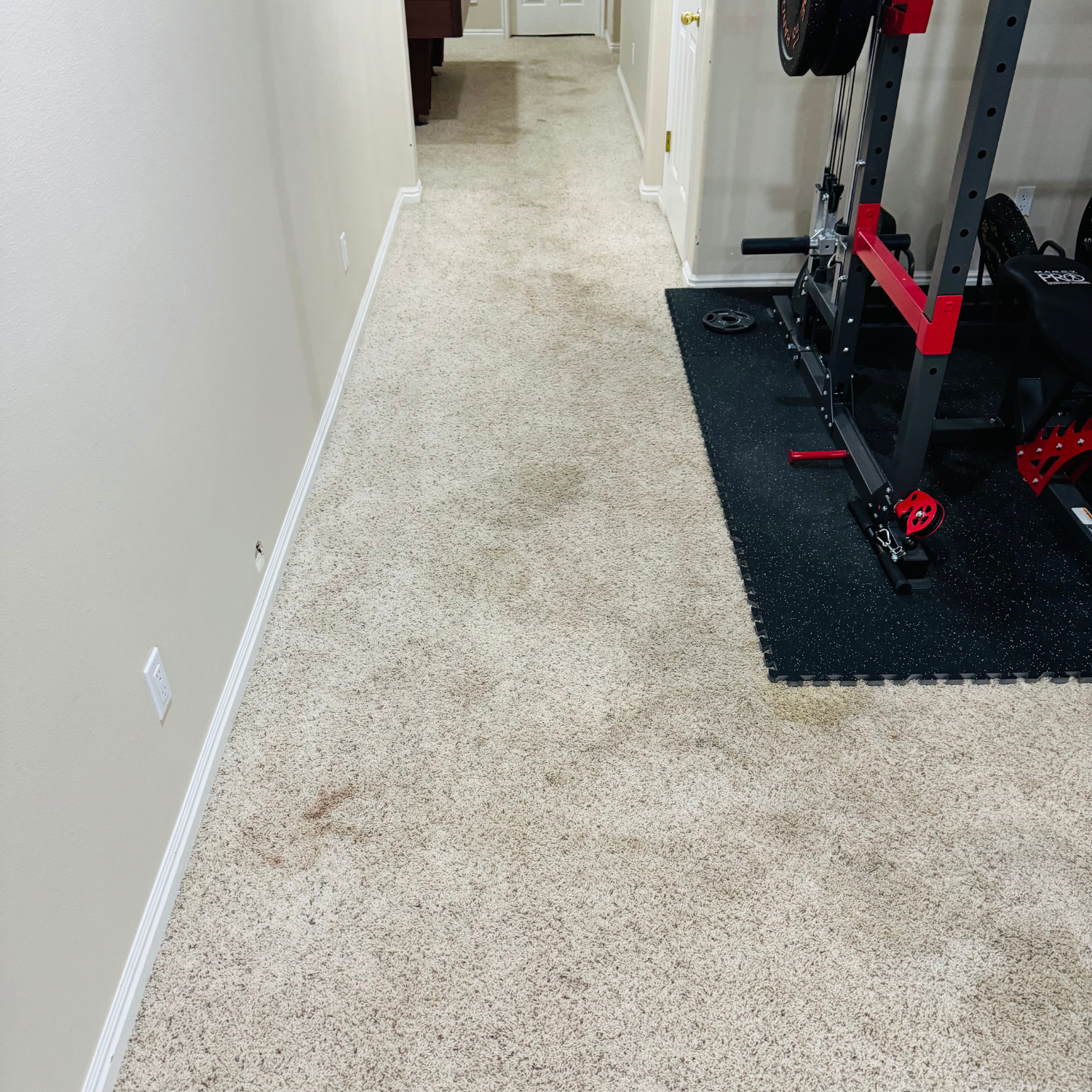 A hallway with beige carpet leads past a gym rack on a black floor mat to a door in the distance.
