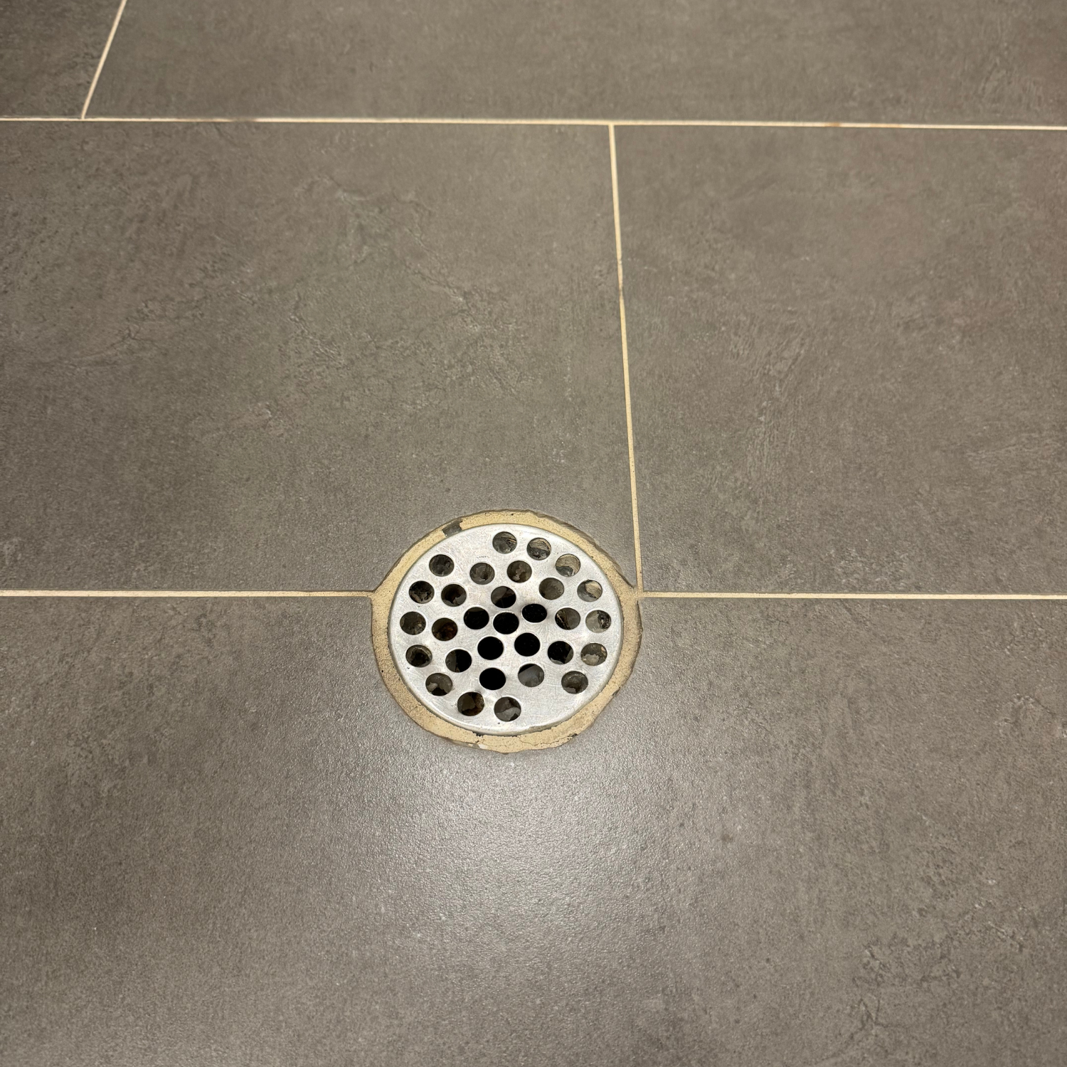 A round metal floor drain cover set into grey tiled flooring.