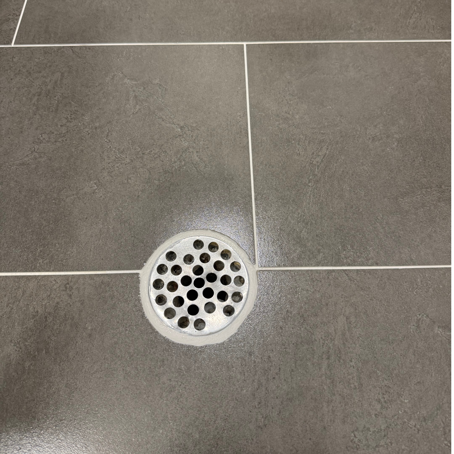 A circular metal shower drain cover set into grey, square-tiled flooring with white grout lines.