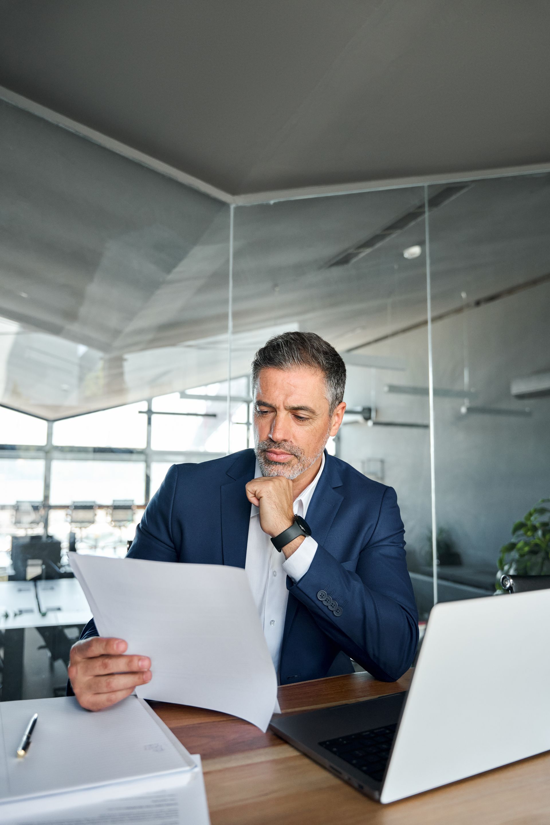 Man in blue suit reviewing a document at a desk with a laptop in a modern office