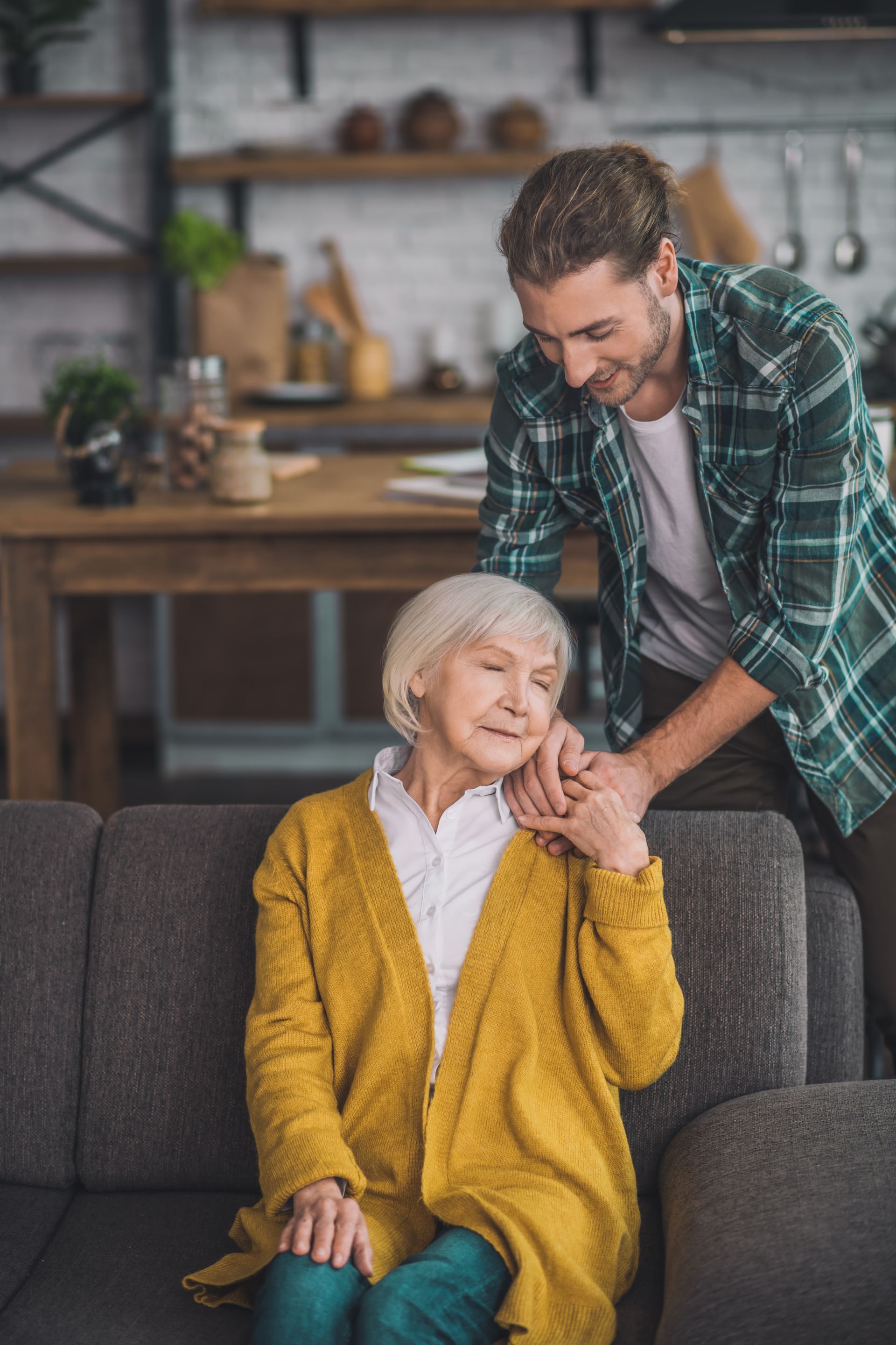 Elderly woman sitting on a sofa while a man in a green plaid shirt gently massages her shoulder
