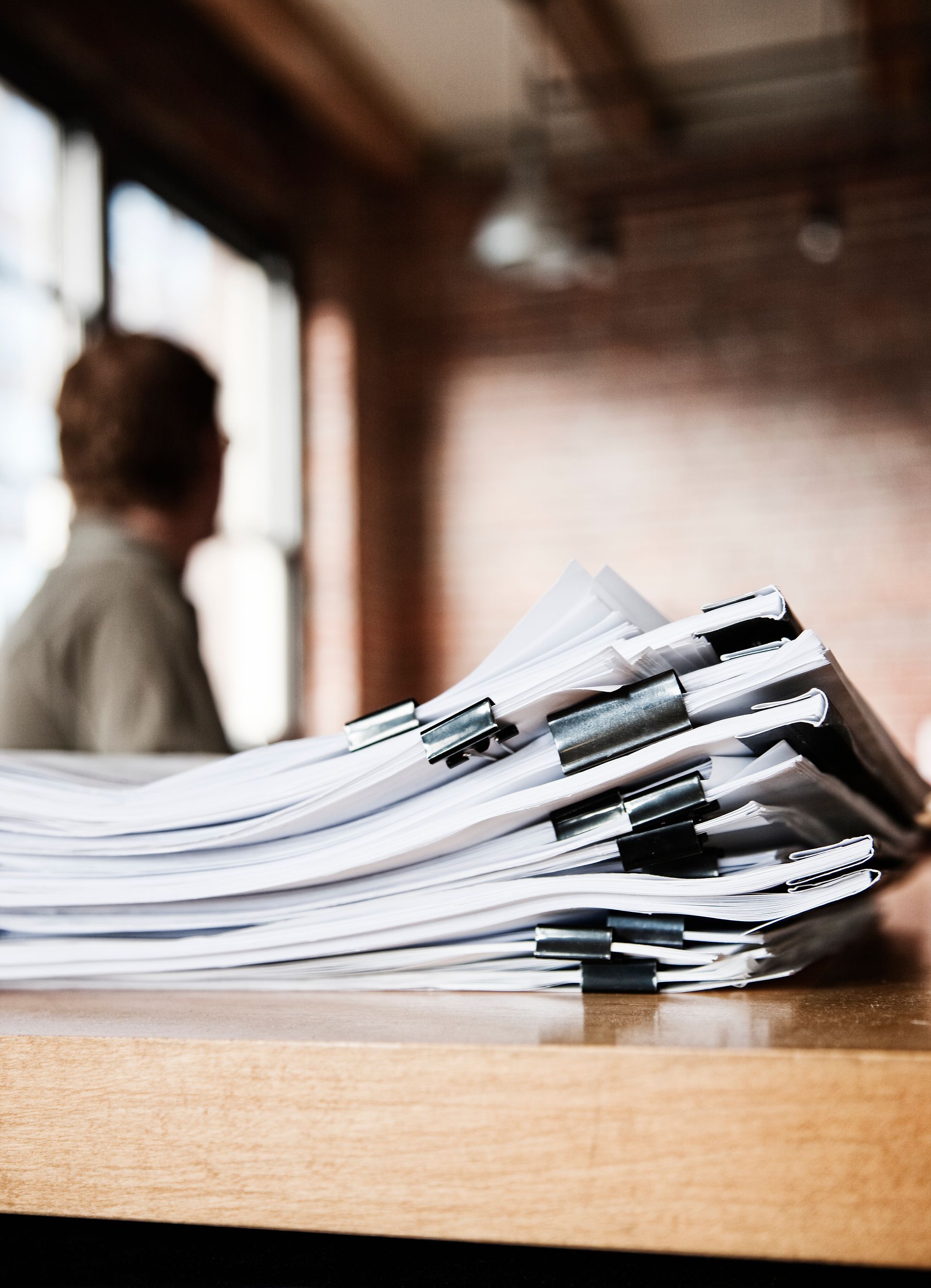 Stack of papers on a wooden table with a blurred person in the background in an office setting