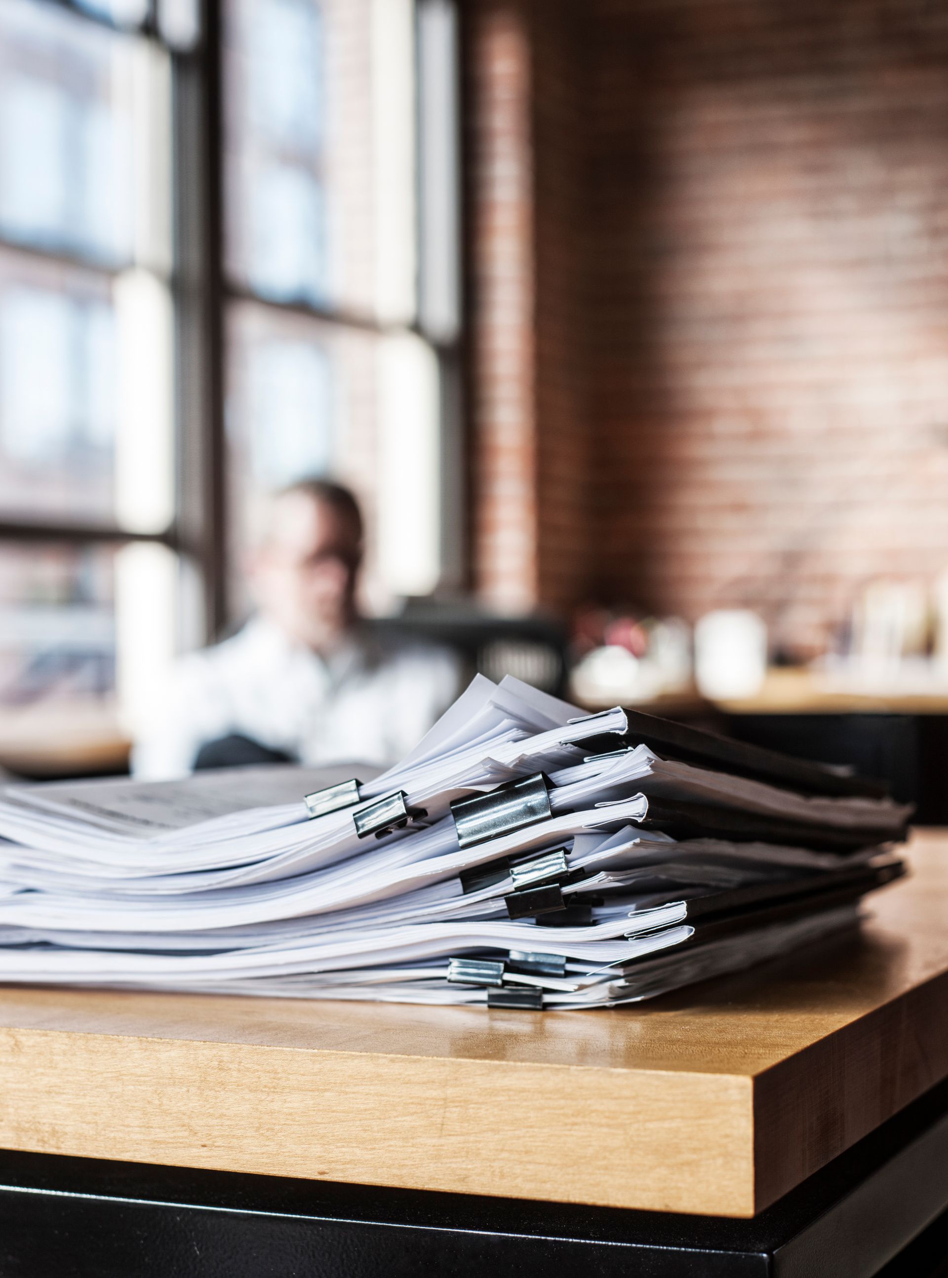 Stack of papers on a wooden table in a bright office, with a blurred person in the background