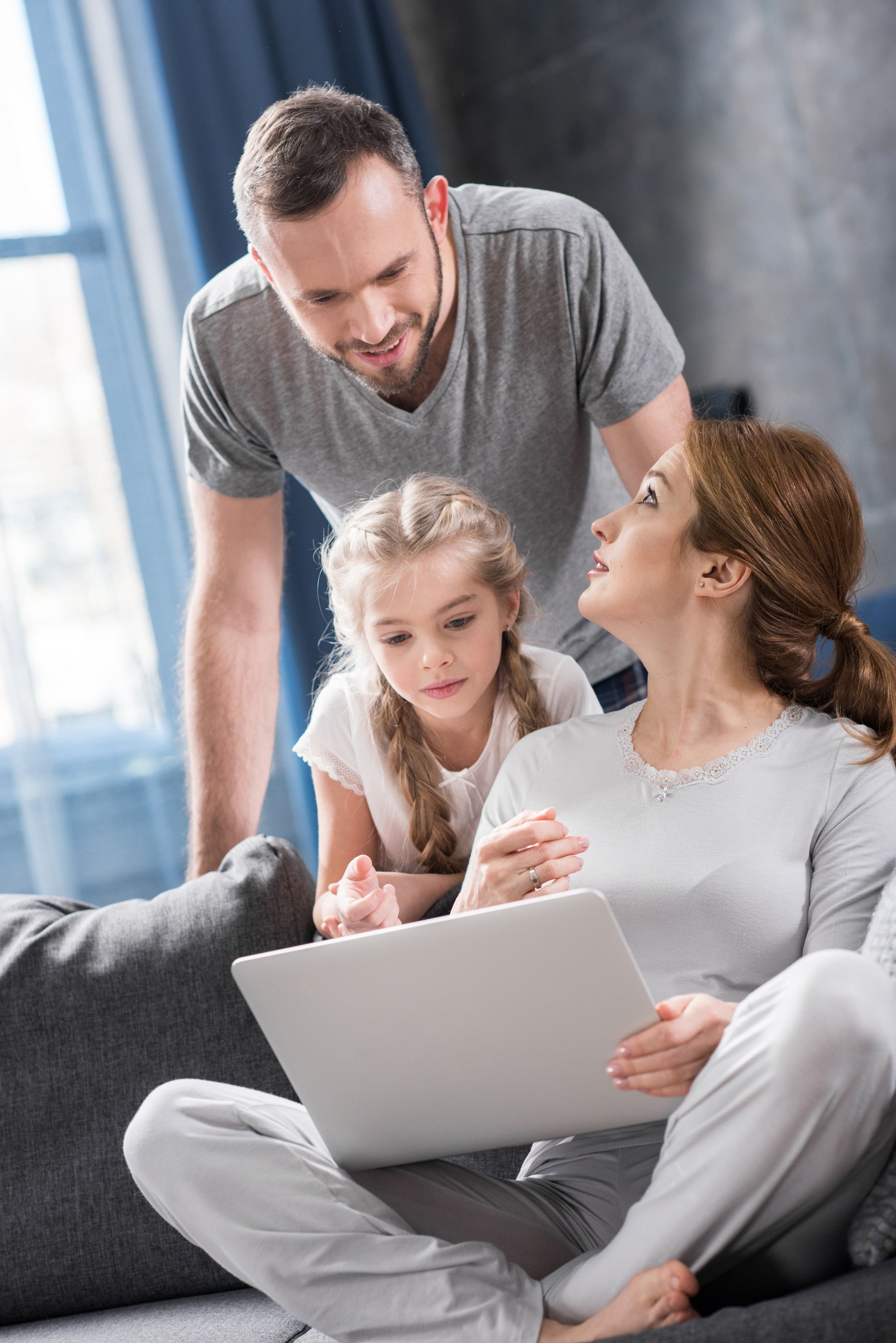 Family gathered around a laptop on a couch, smiling and talking in a bright living room.