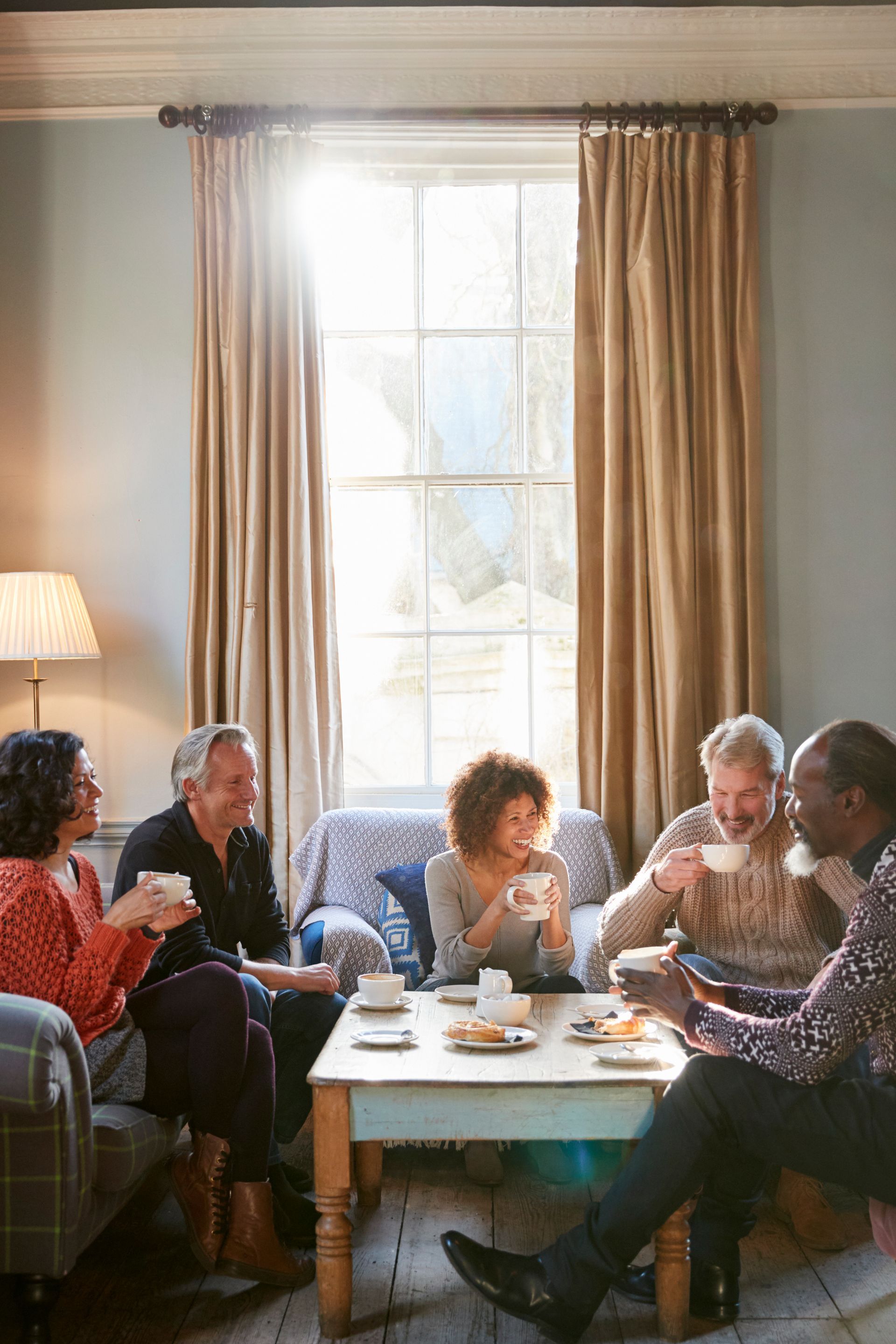 Five people sitting around a coffee table in a sunlit living room, sharing a meal and talking