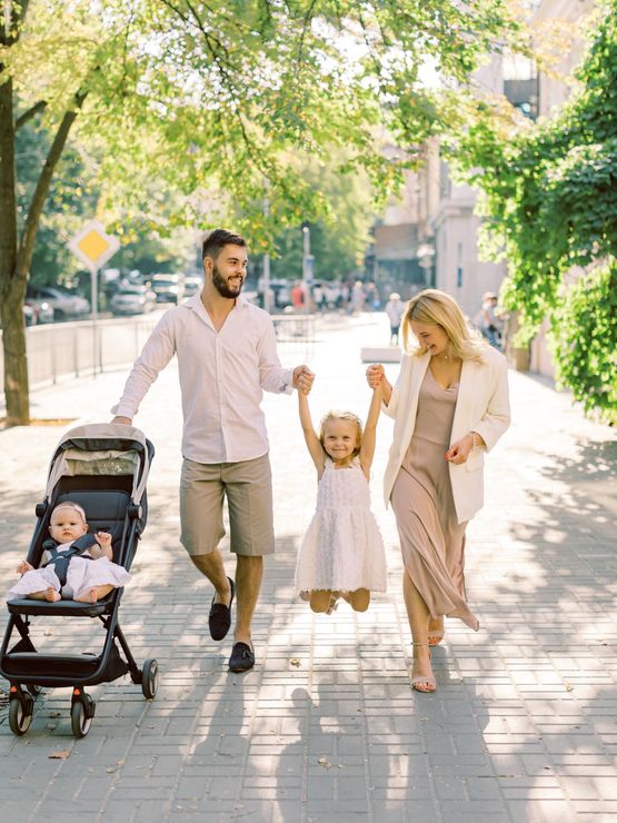 Family walking on a sunny tree-lined sidewalk, with a stroller and two children holding hands
