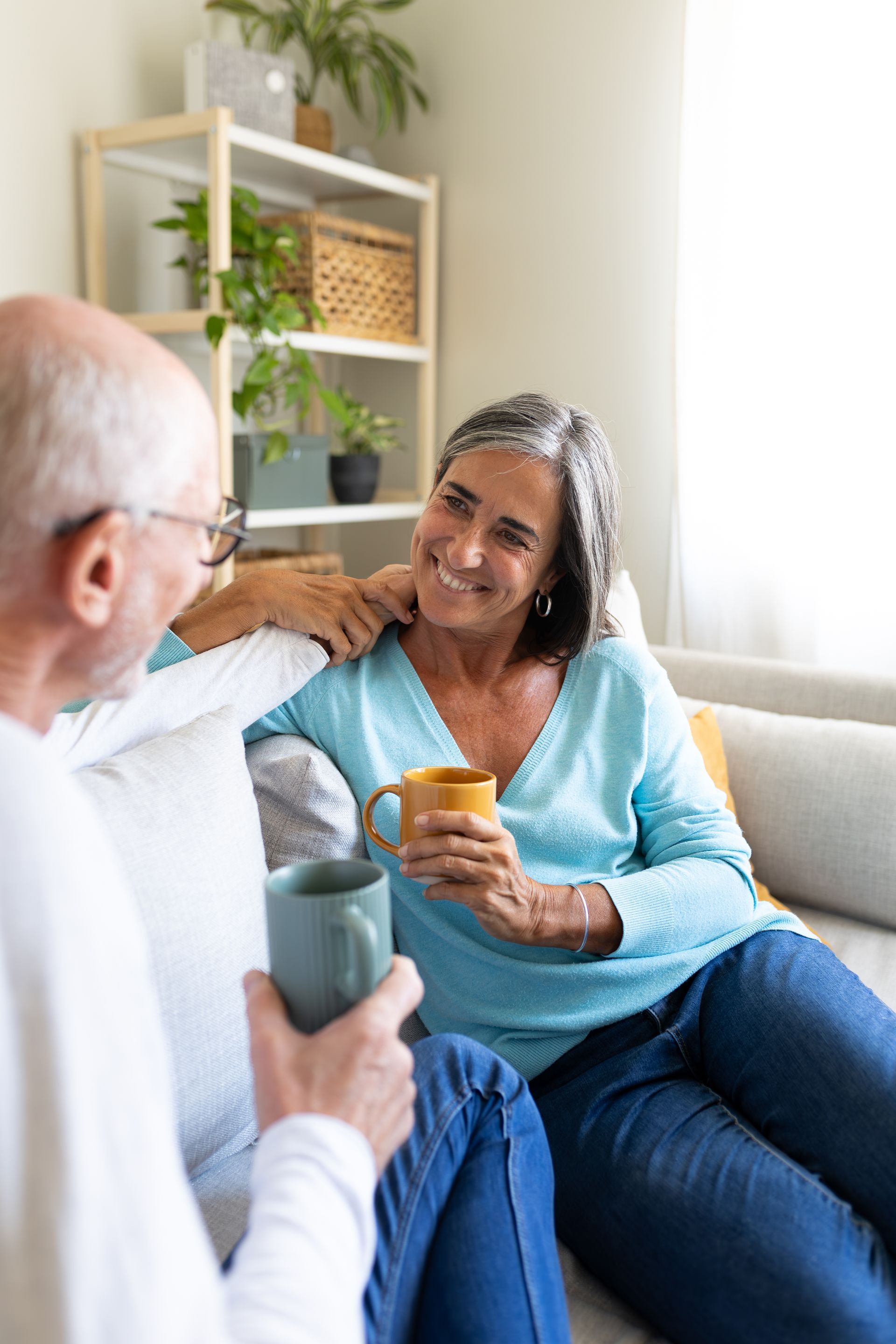 Two people sit on a couch talking and smiling while holding mugs in a bright living room.