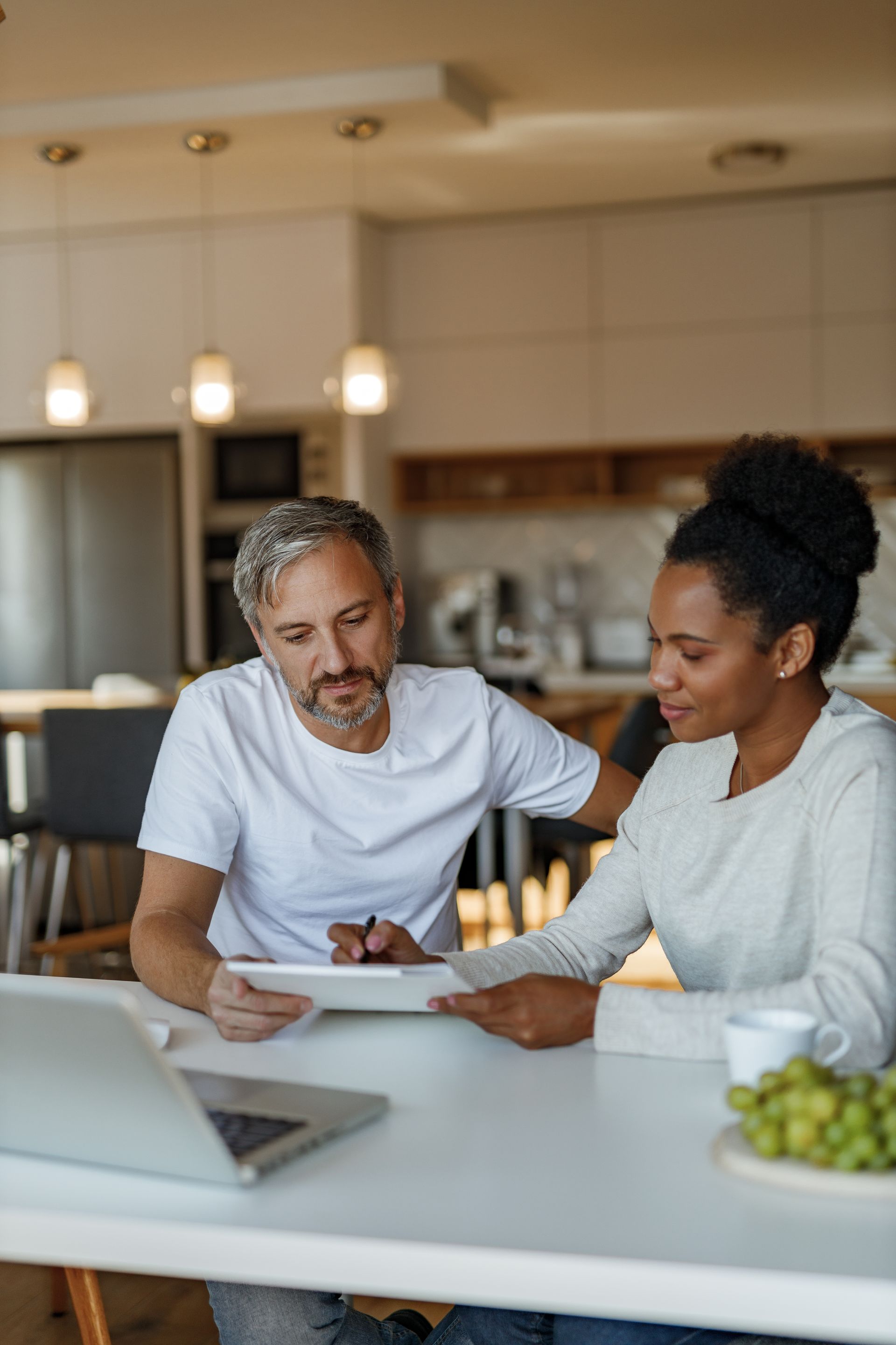Two people reviewing documents at a kitchen counter with a laptop and coffee mugs