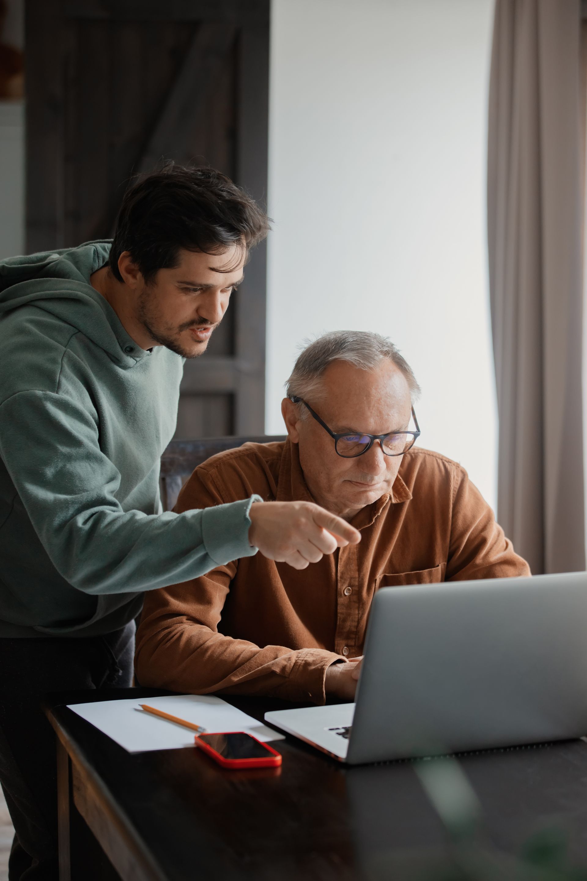 Two people reviewing documents beside a laptop at a desk by a window