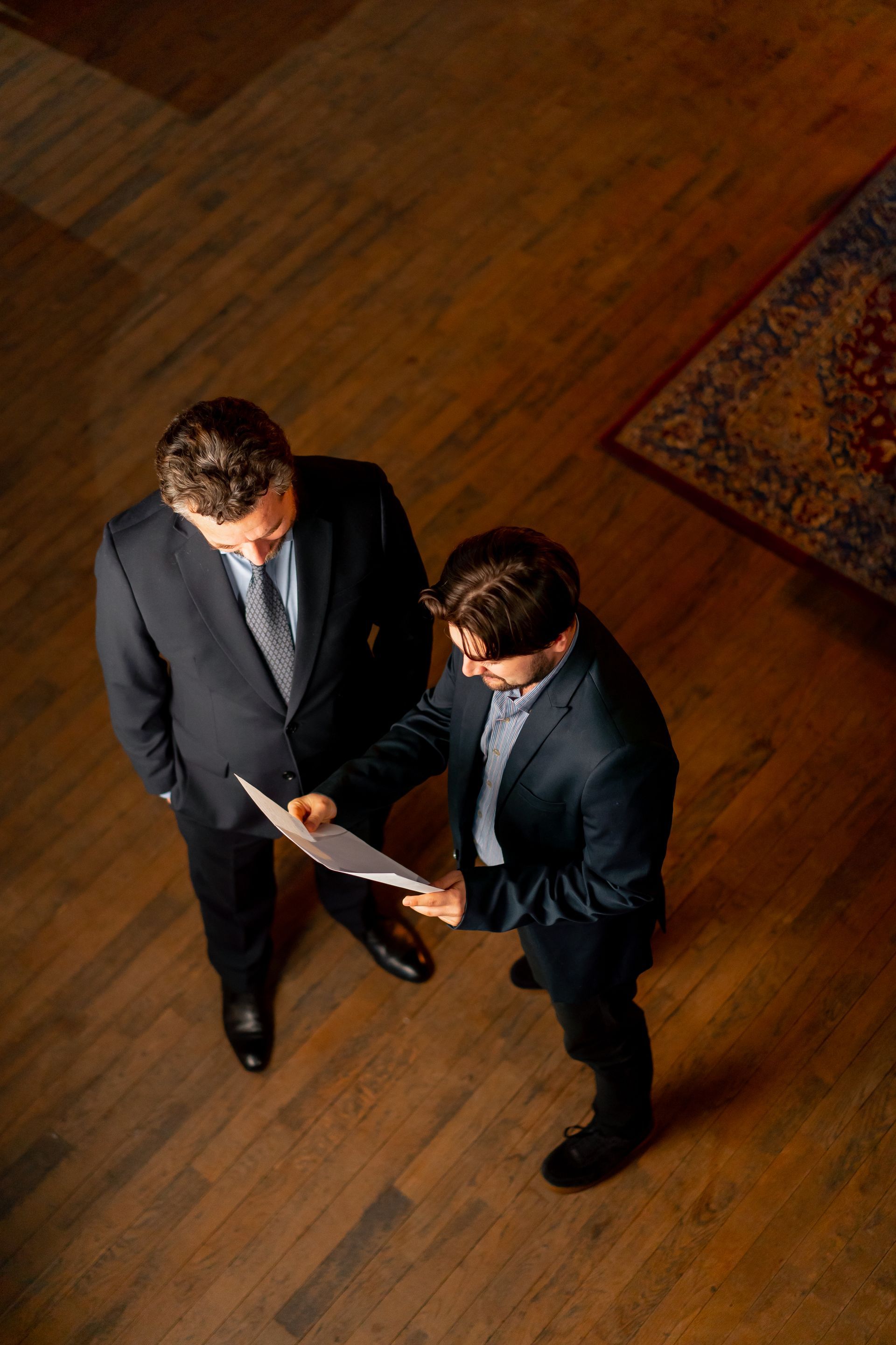 Two people in dark suits reviewing a document on a wooden floor, viewed from above.