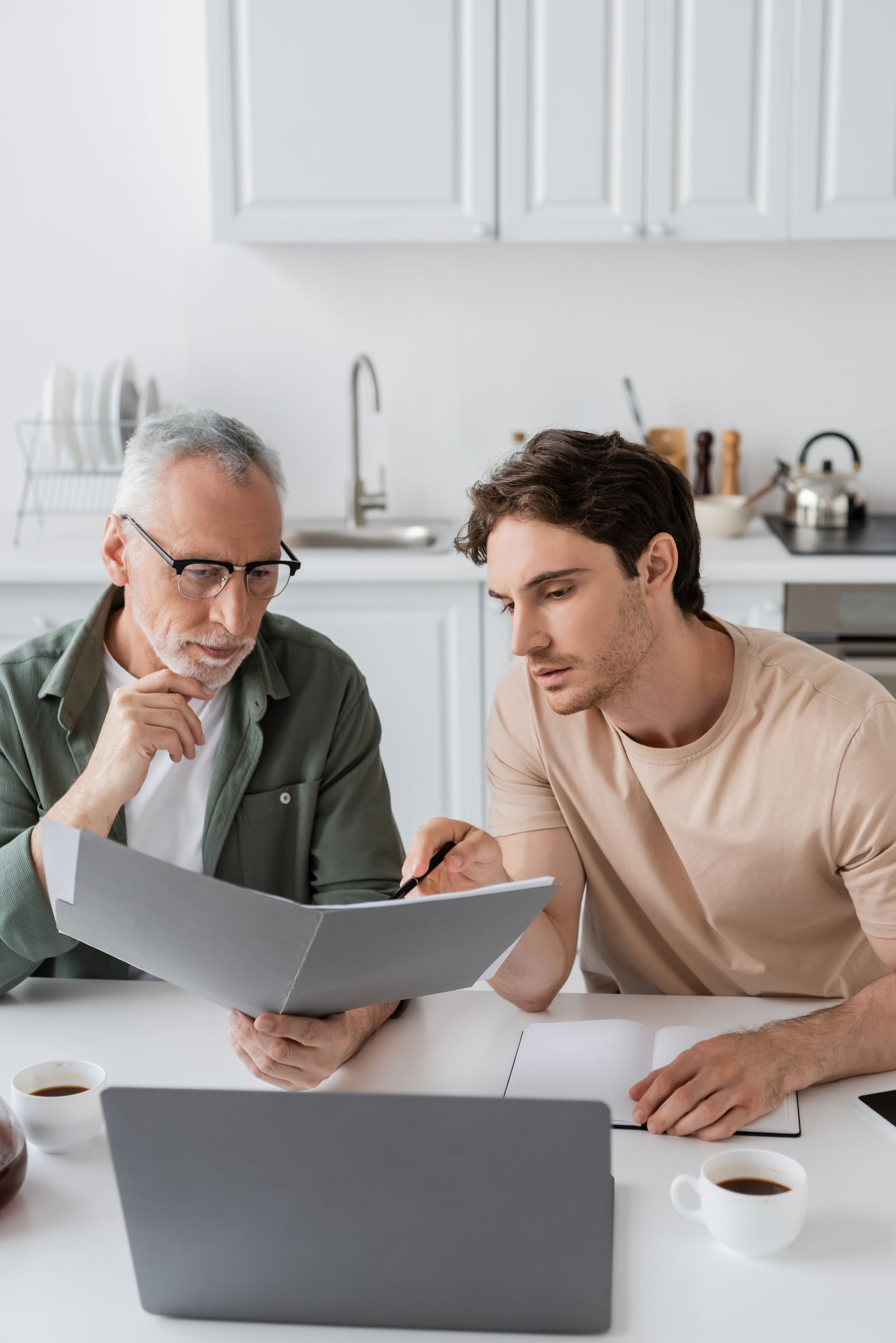 Two people reviewing documents together at a white kitchen table with a laptop and coffee cups