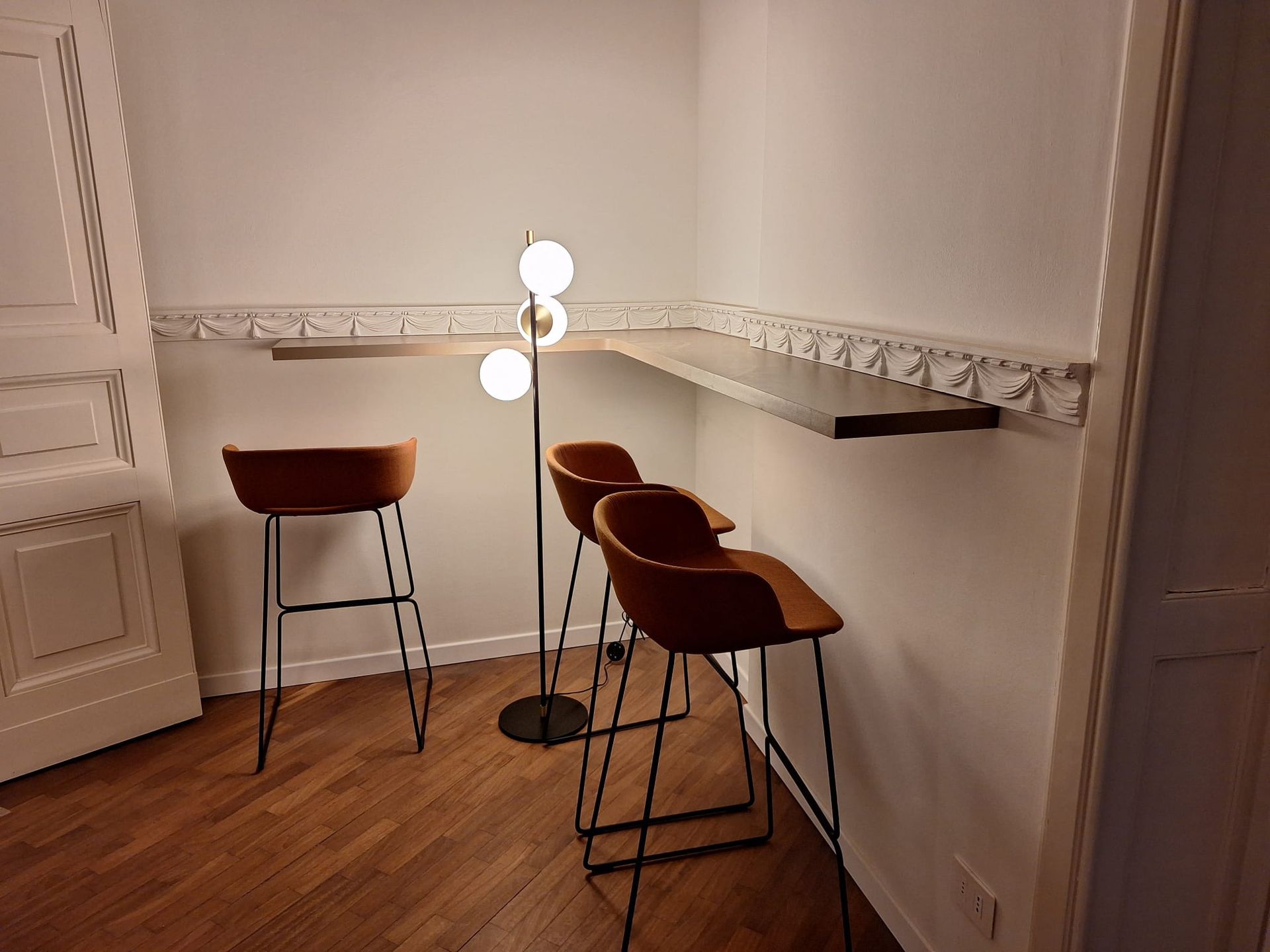 Moderno angolo di stanza con pareti bianche, pavimento in legno e tre sgabelli da bar marroni accanto a una lampada da terra.