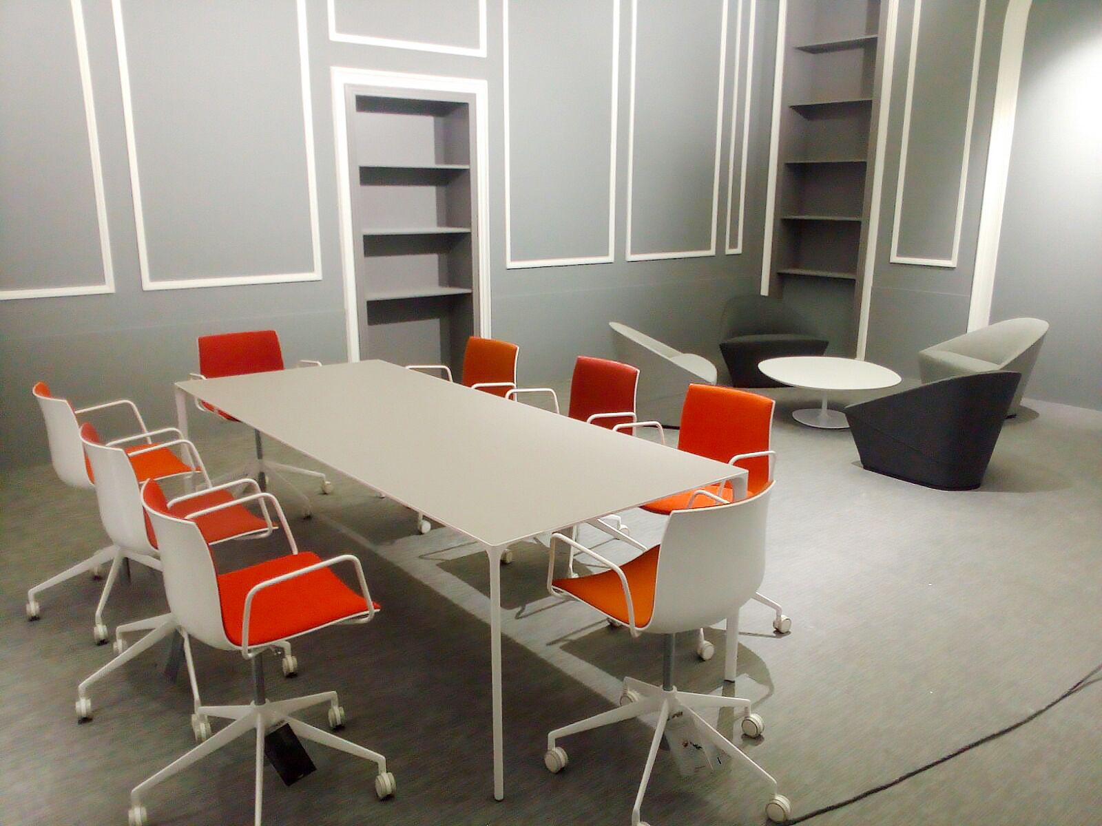 Moderna area relax per ufficio con sedie arancioni attorno a un tavolo bianco e zona salotto grigia.