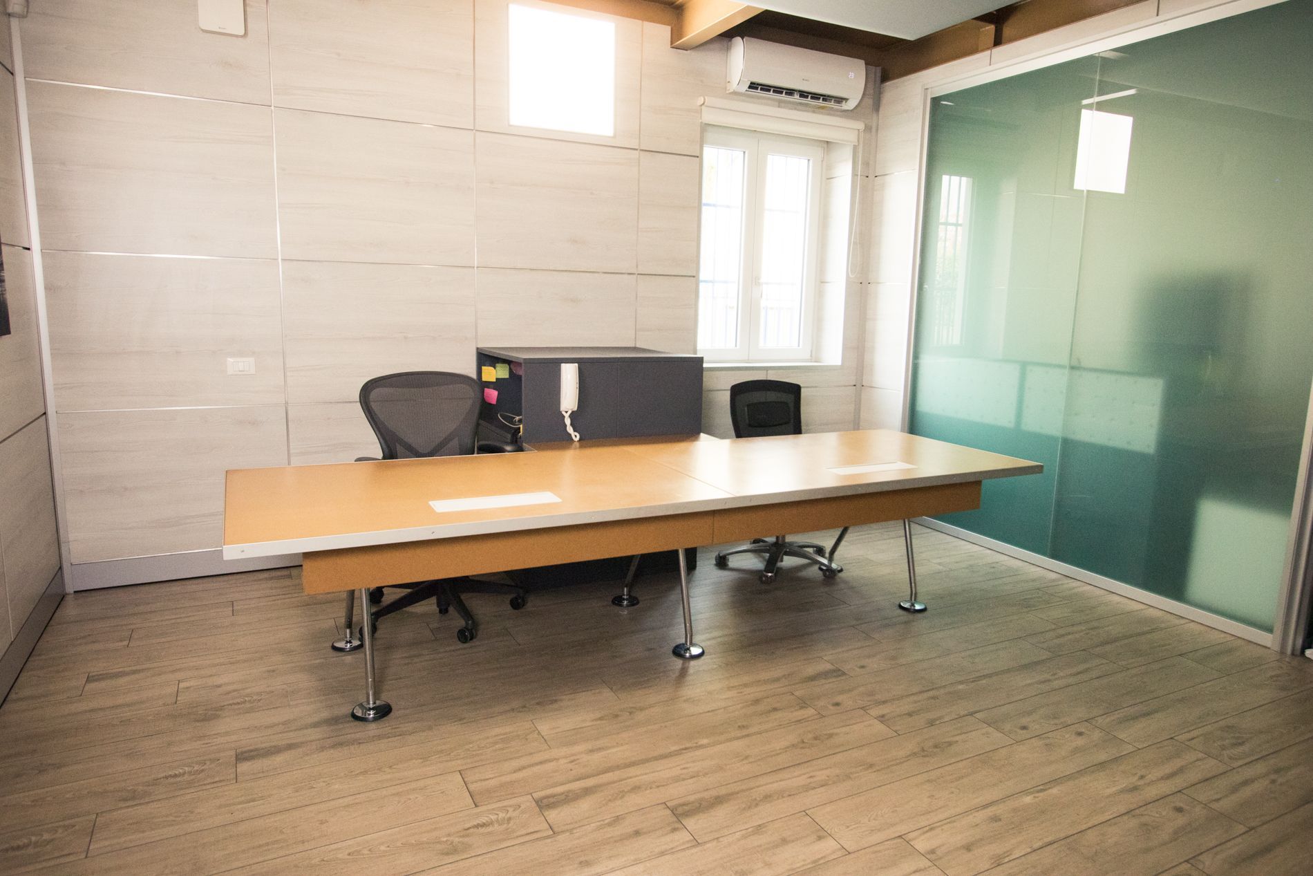 Sala riunioni vuota in ufficio con lungo tavolo di legno, sedie, parete in vetro e finestra luminosa.