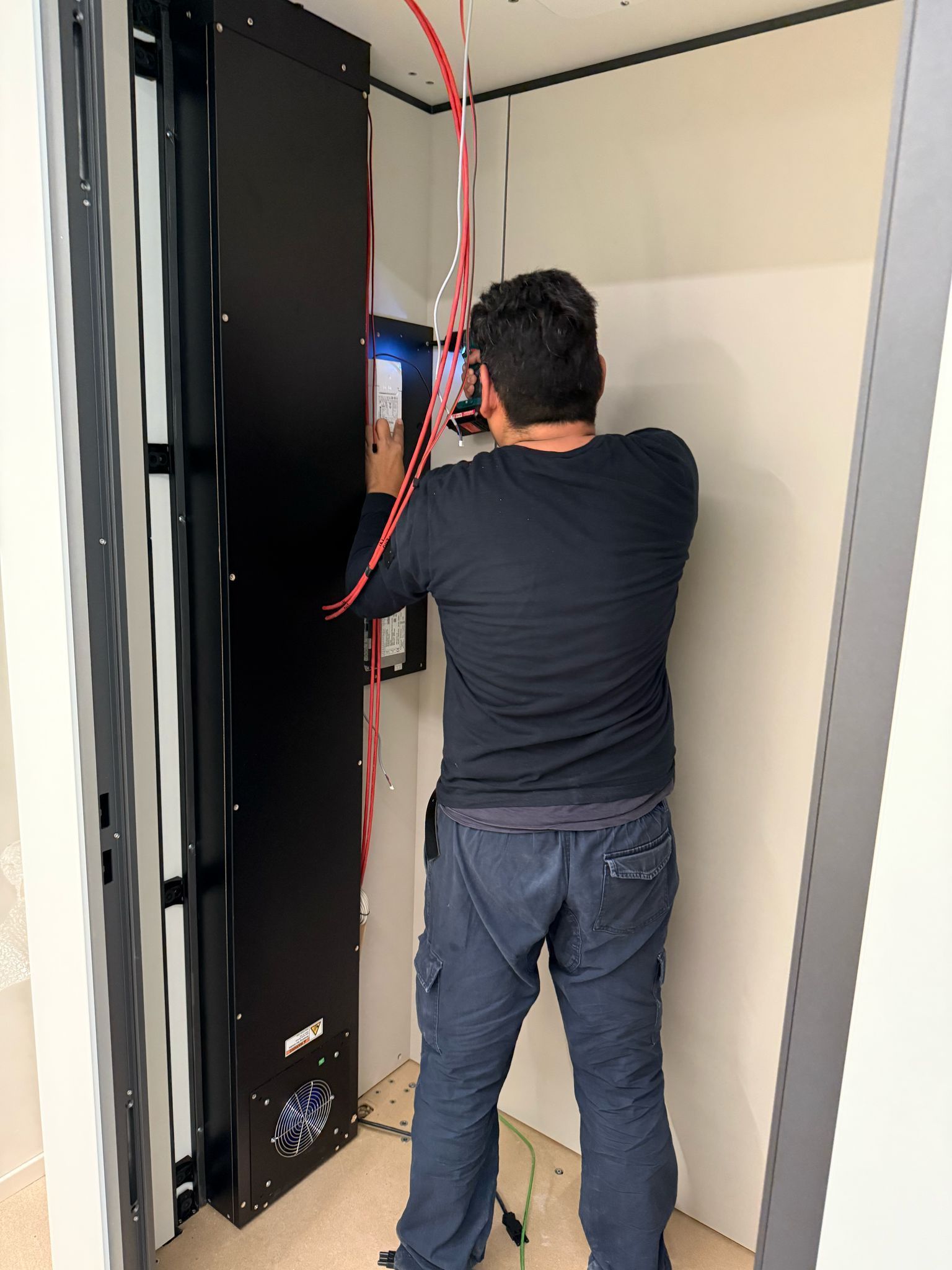 Una persona lavora all'interno di un quadro elettrico aperto, con cavi rossi e un pannello nero in una stanza stretta.