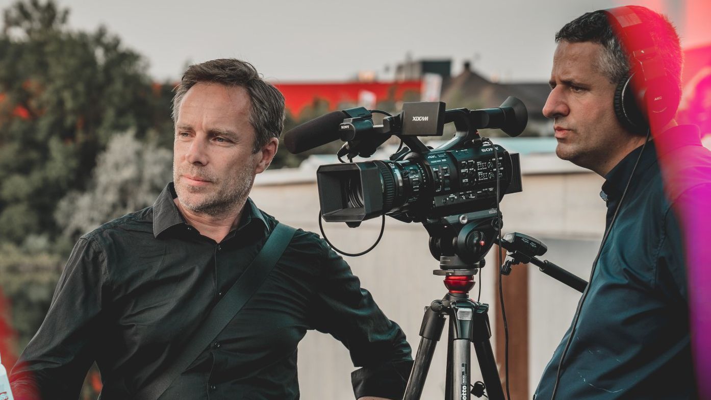 Two men are standing in front of a camera on a tripod.