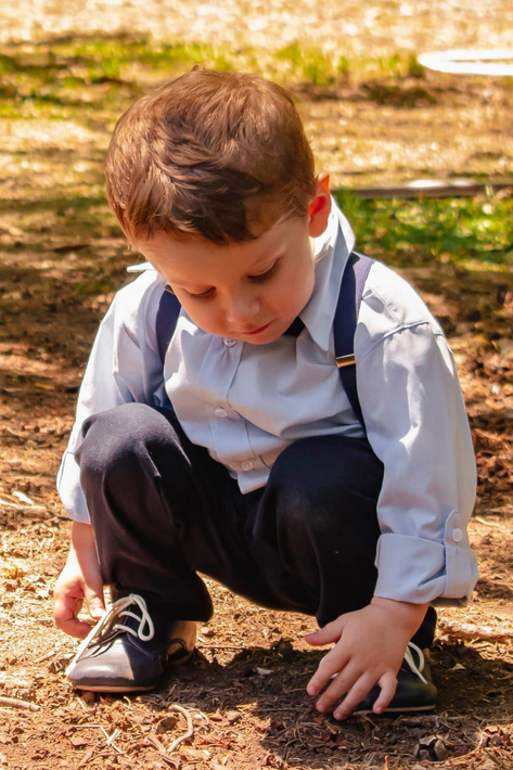 Niño agachado al aire libre, tocando el suelo, vestido con una camisa blanca, pantalones oscuros y tirantes.