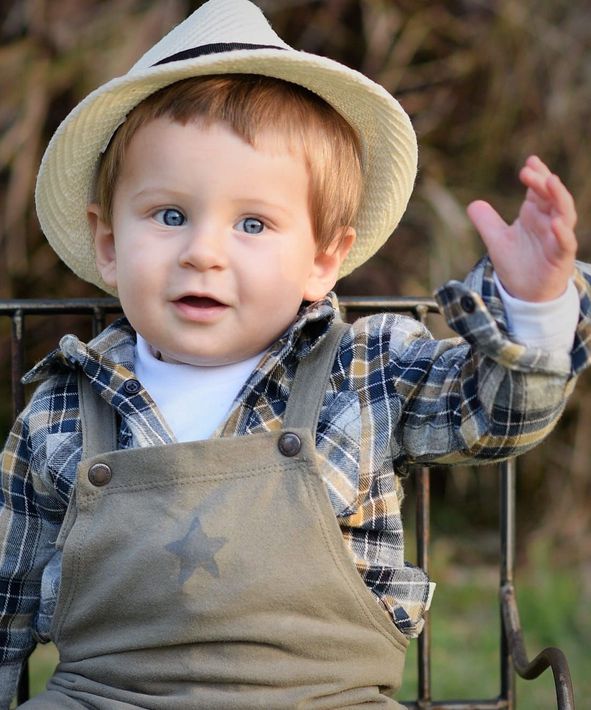 Niño pequeño con sombrero de paja y camisa a cuadros, levantando la mano al aire libre, sonriendo.
