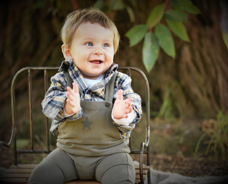 Un niño sonriente aplaude sentado en una silla al aire libre, vestido con una camisa a cuadros y un mono.
