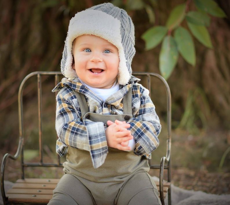 Un bebé sonriente, con gorro gris y camisa a cuadros, está sentado en un banco al aire libre con las manos entrelazadas.