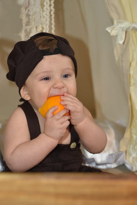 Un bebé con un conjunto y gorro negros, mordisqueando un juguete naranja, está sentado en el interior de una casa .
