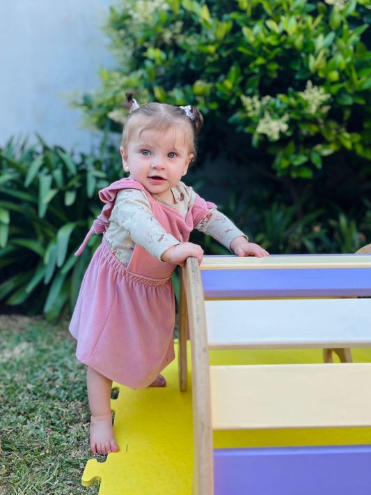 Una niña pequeña con un vestido rosa se sube a un banco amarillo del parque infantil y mira a la cámara.